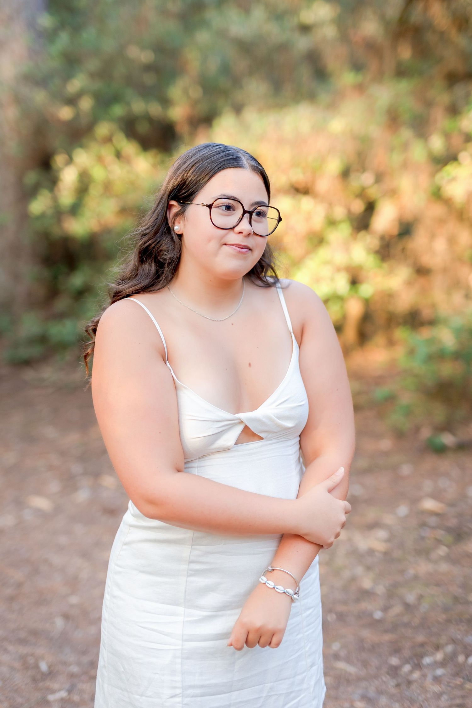 A portrait of someone in a white dress with glasses standing on a forest path.