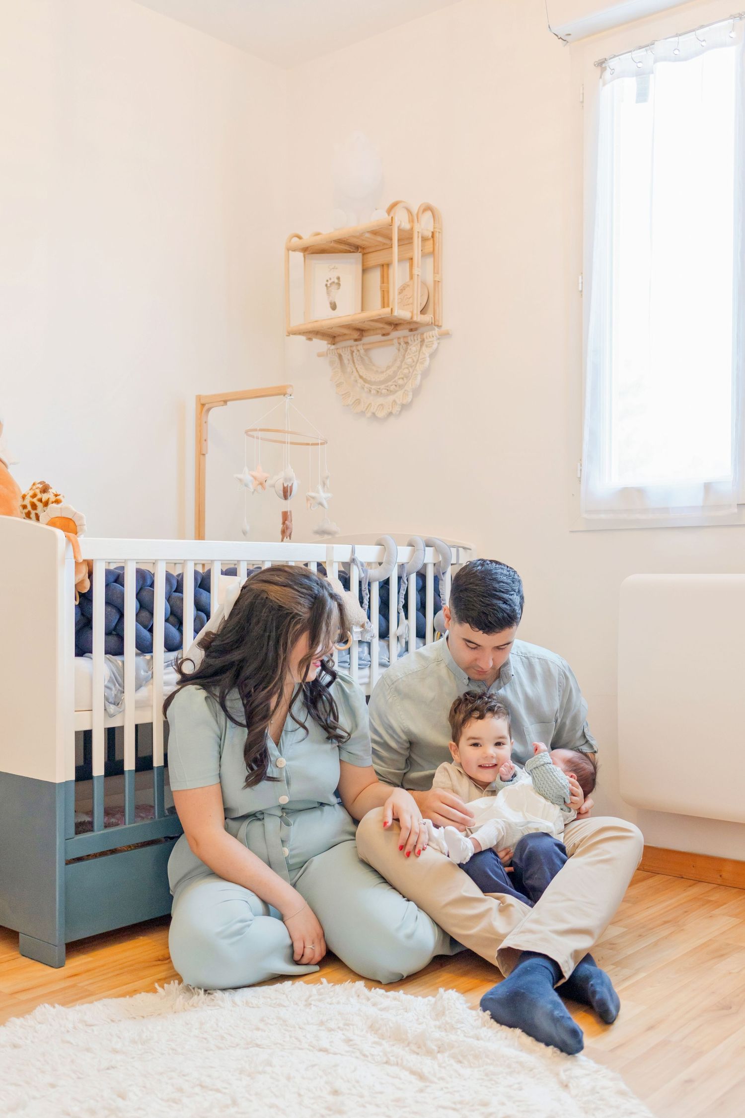 Family sitting together on white rug in nursery with blue crib and wooden shelving.