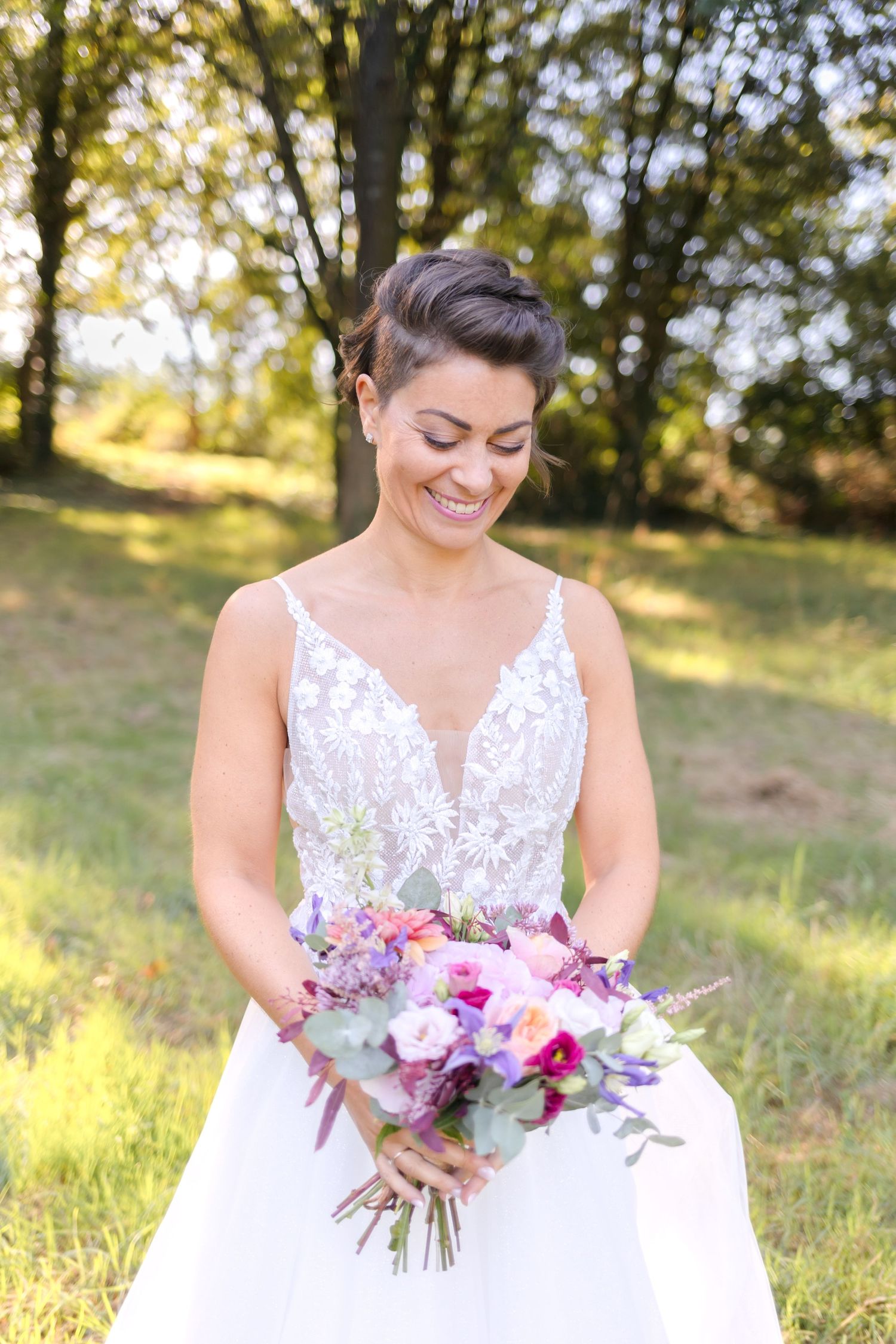 A bride in a white lace dress holds a vibrant purple and pink bouquet in sunlit garden setting.