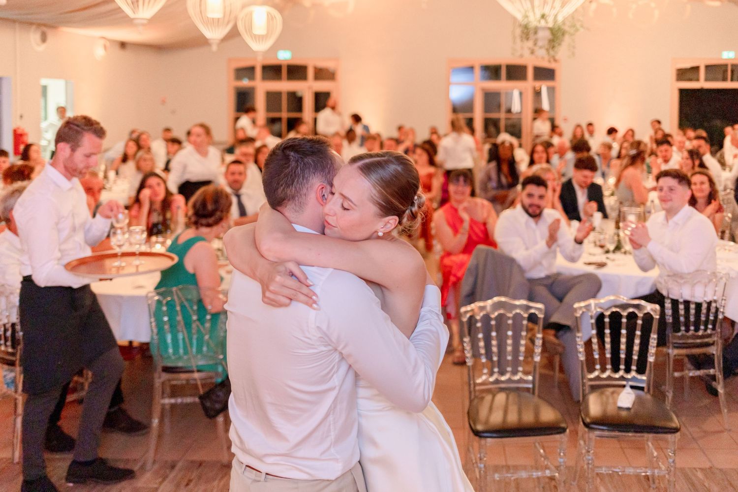 Wedding couple embraces during their first dance as guests watch from tables in a beautifully lit reception venue.