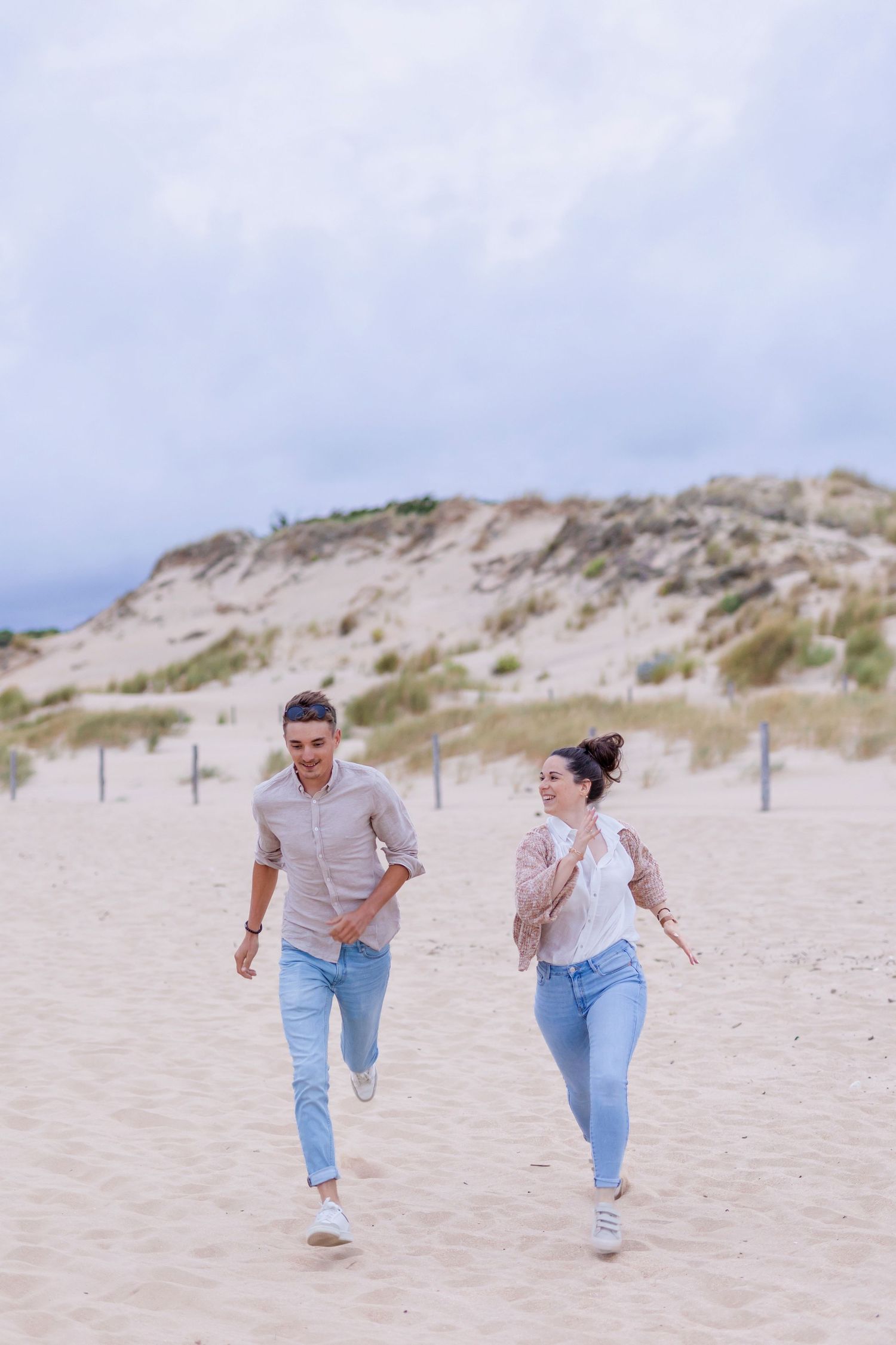 Playful moment of two people running on sandy beach with dunes in background.