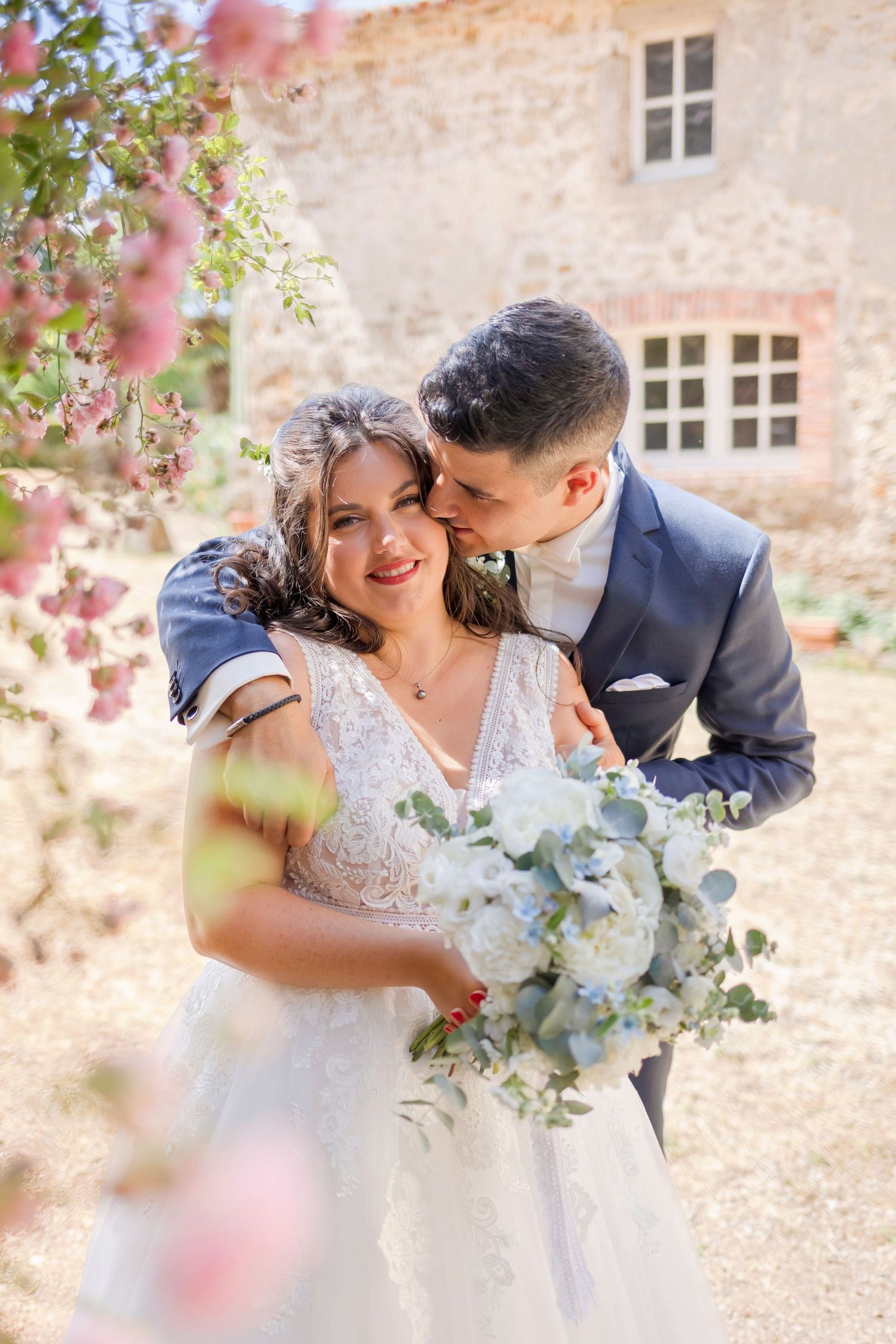 Wedding couple shares romantic moment among pink flowers outside rustic stone building.