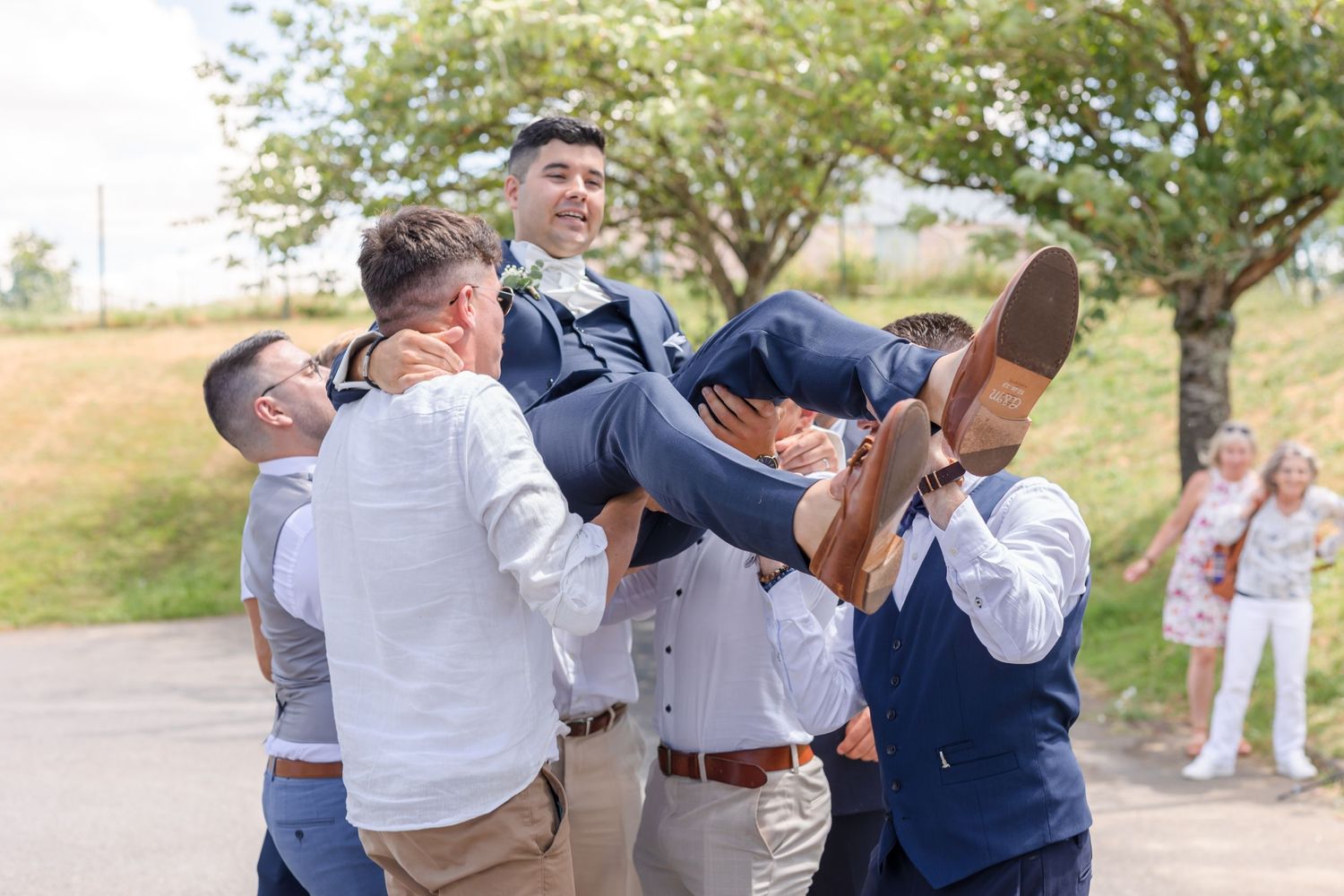 Wedding party groomsmen lifting someone up in celebratory moment outdoors.