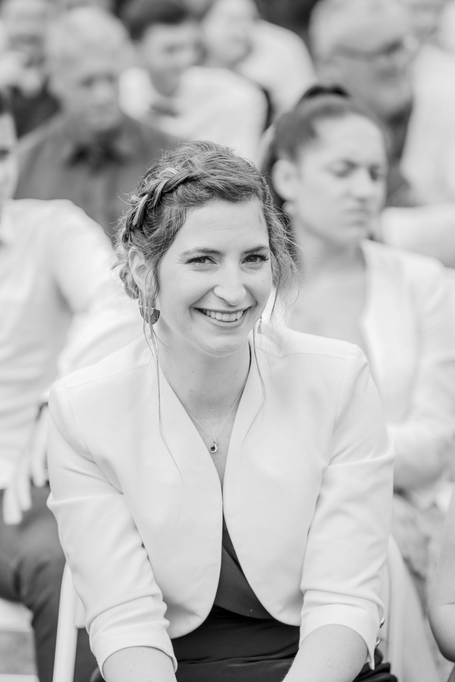 A guest in a white blazer smiles during an outdoor wedding ceremony.