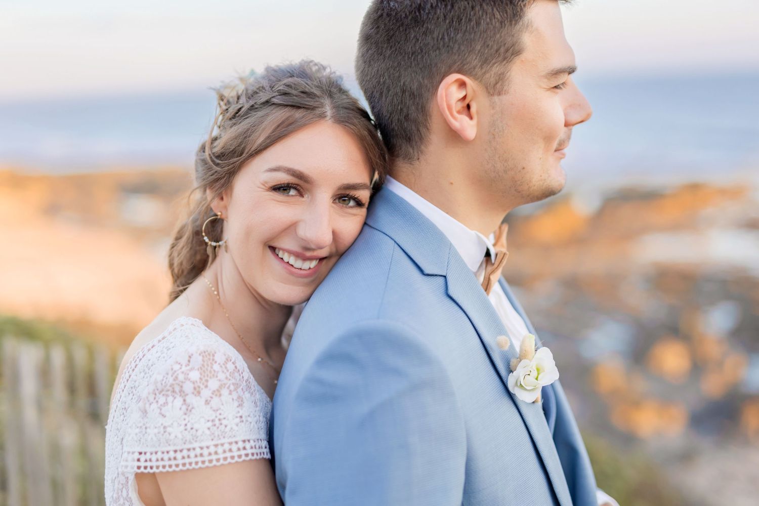 Wedding couple in blue suit and white dress posing together against coastal sunset backdrop.