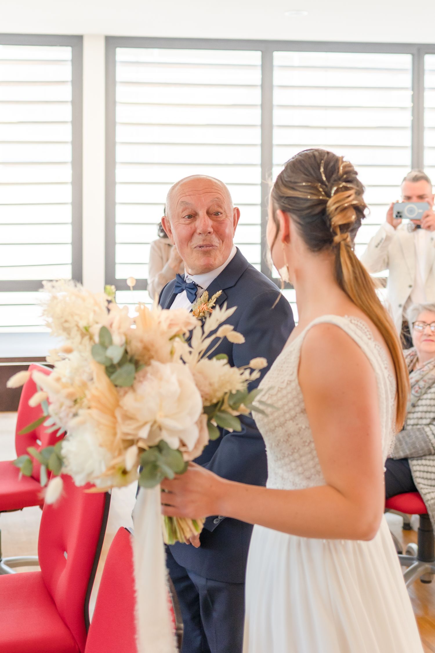 Wedding ceremony attendees gather around an elegant floral display on red chairs.