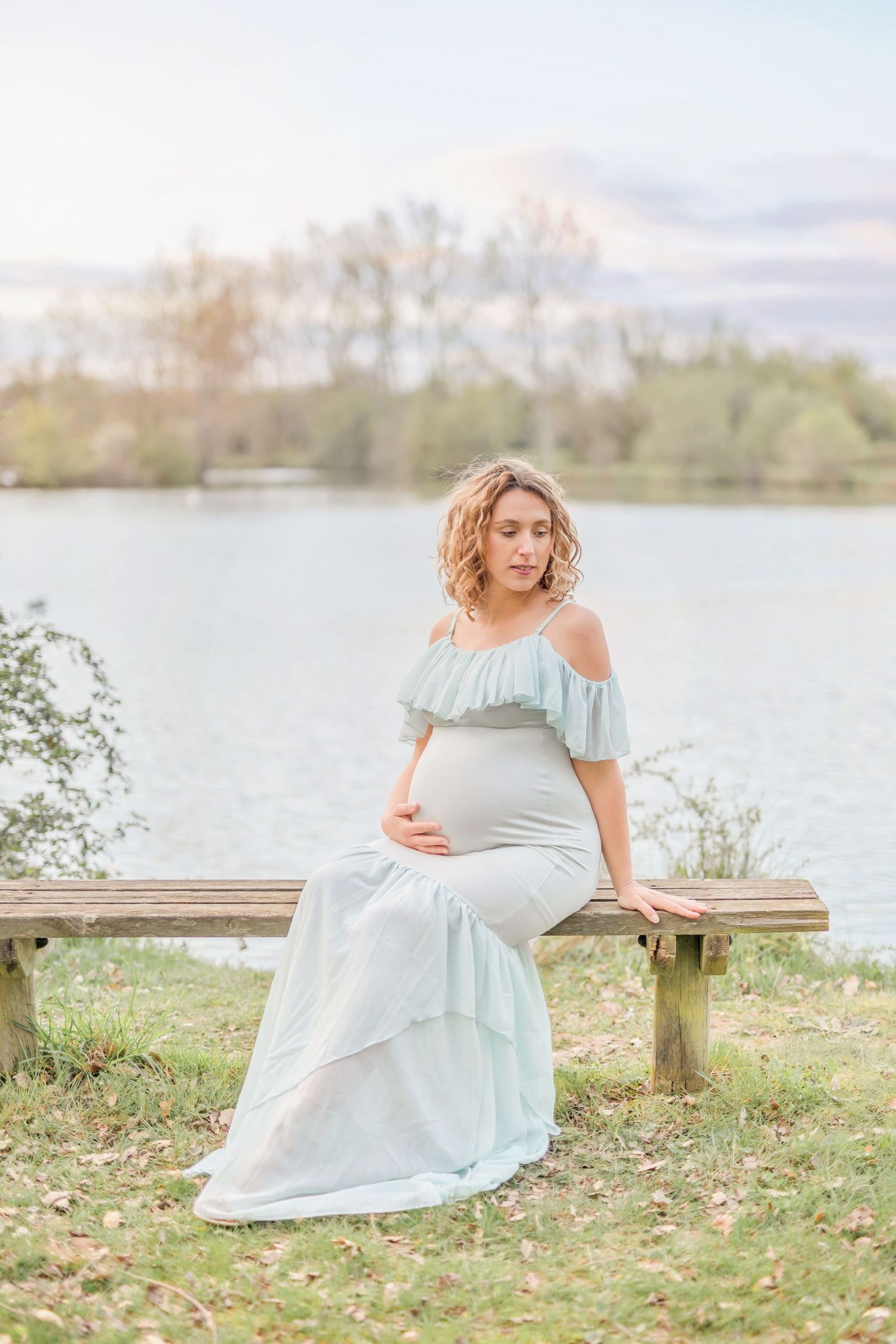 Romantic maternity photoshoot in a flowing white dress by a serene lakeside setting.