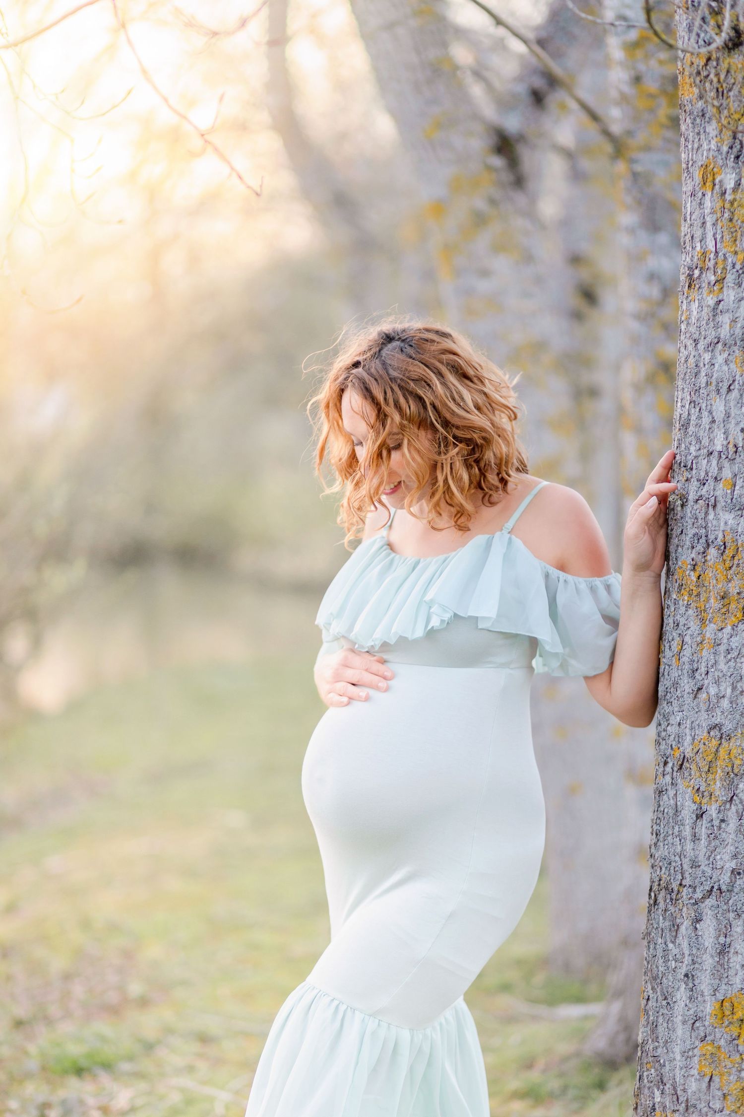 Artistic maternity photo leaning against a tree trunk in soft natural lighting.