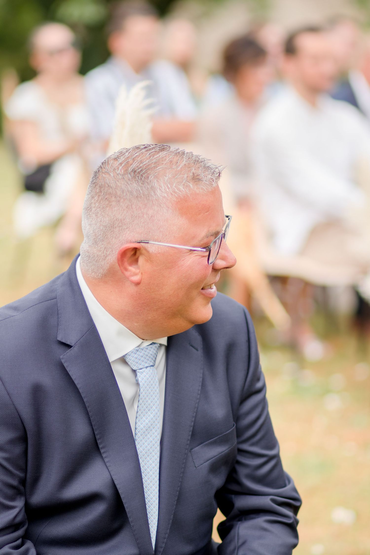 A guest in a navy suit and light blue tie smiles during an outdoor wedding.
