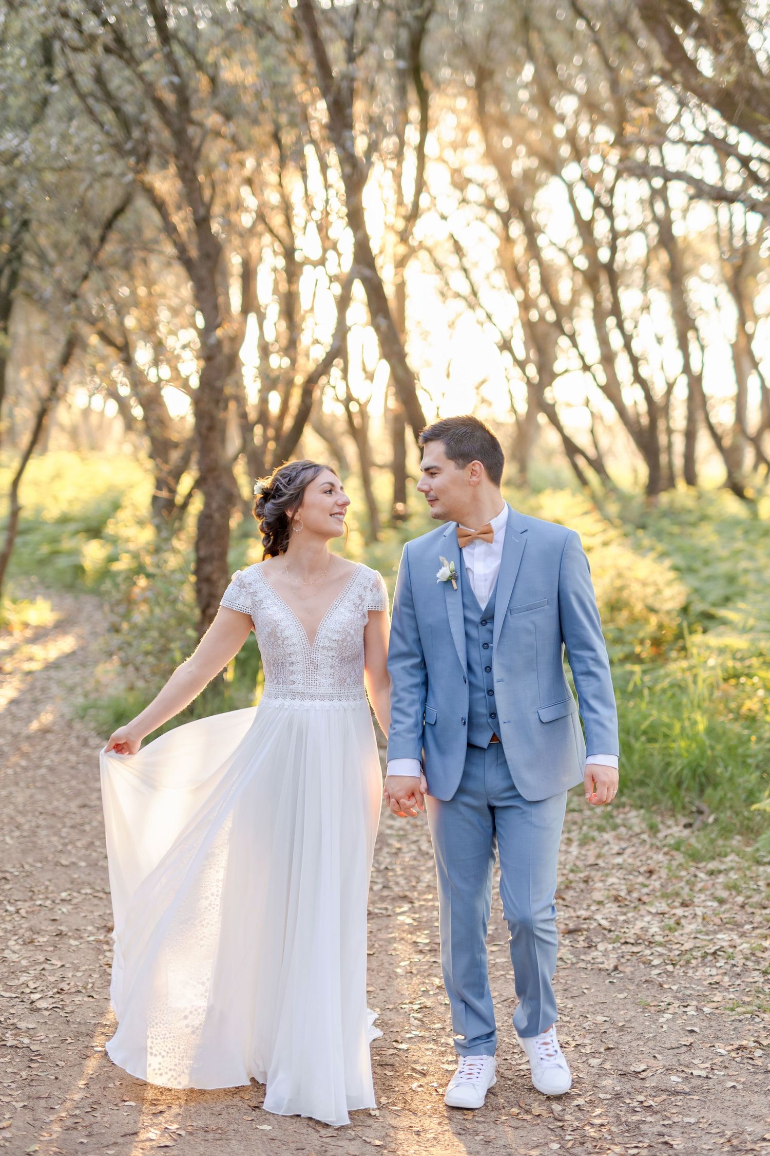 A joyful wedding couple walking hand in hand on a sunlit forest path.