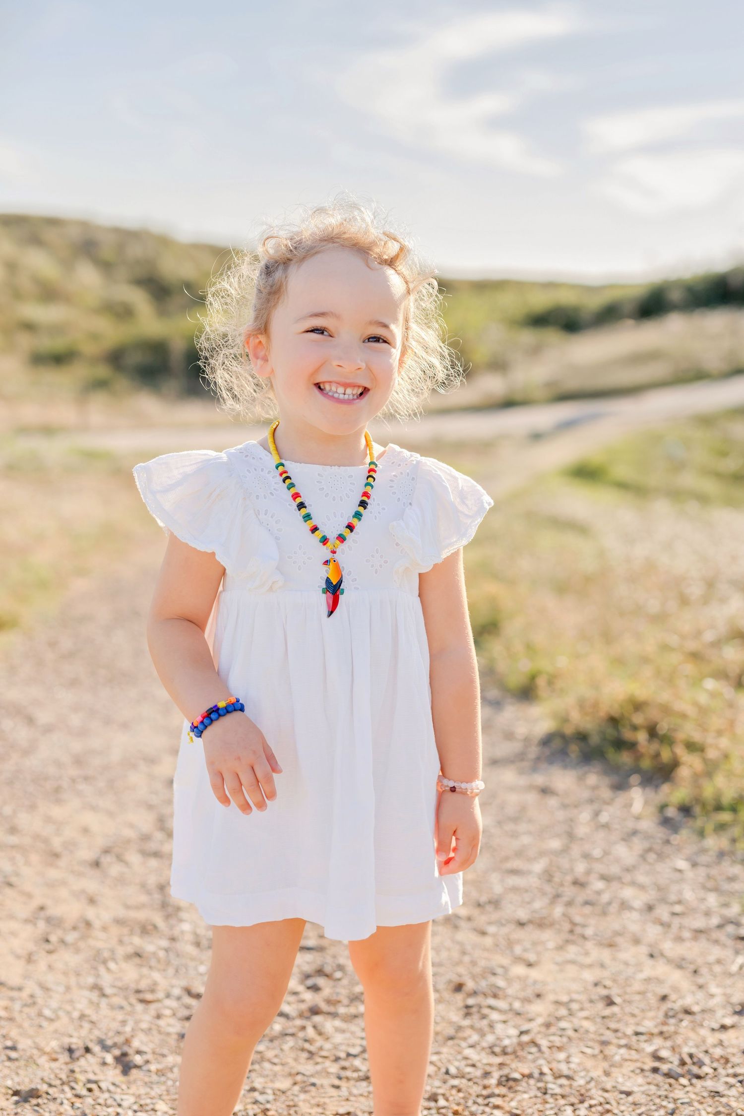 Child in white dress with colorful beaded necklace standing on dirt path in sunlit countryside.
