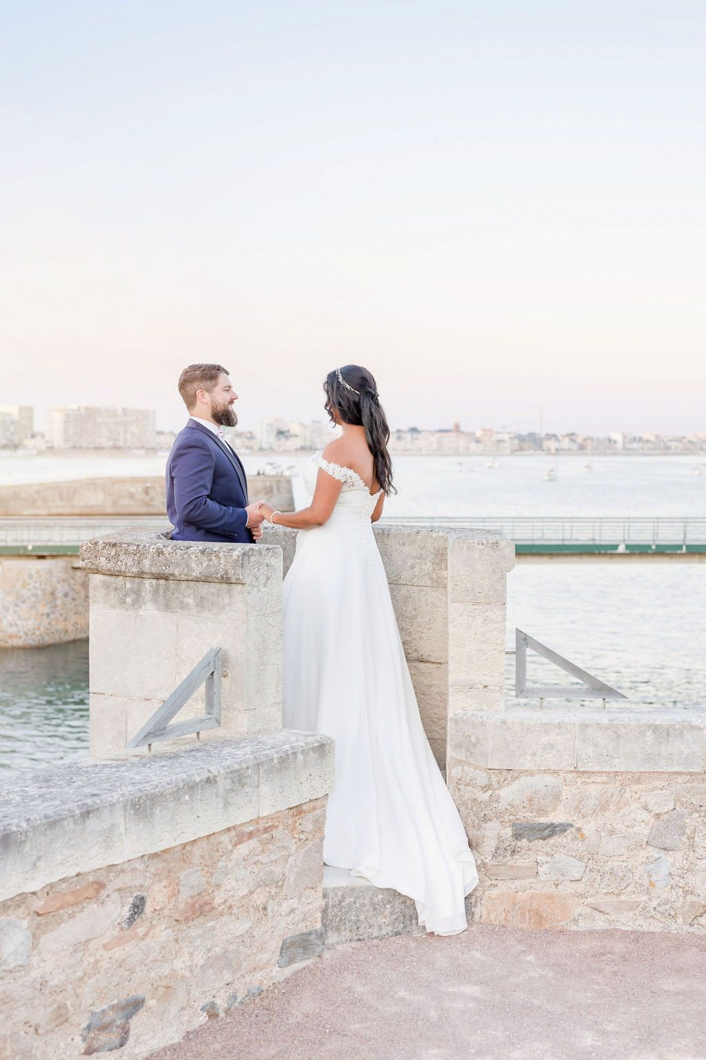 Bride and groom sit together on stone wall overlooking harbor during sunset.