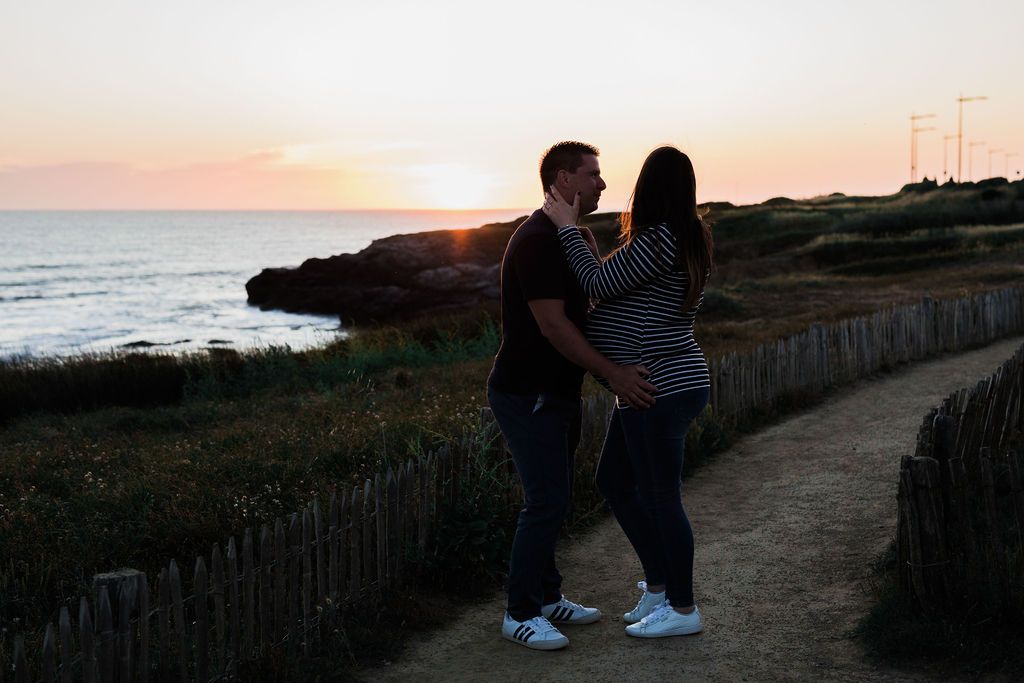 A couple embraces at sunset along a coastal path with ocean views and wooden fence.
