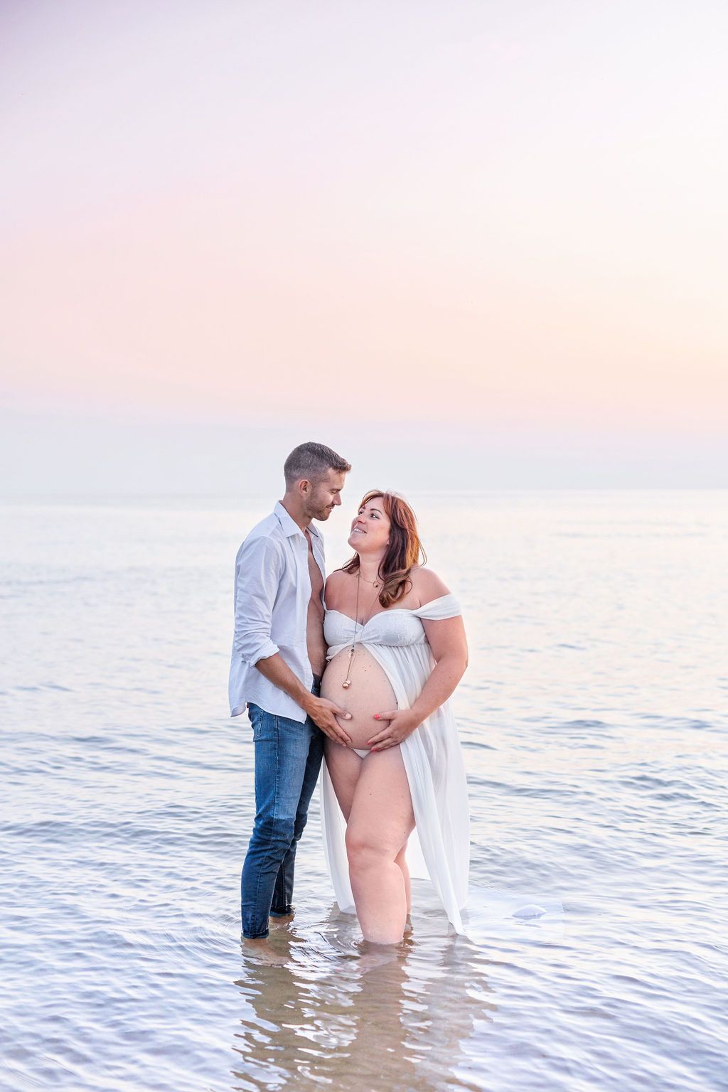 Maternity beach photo session at sunset with couple standing in gentle waves.