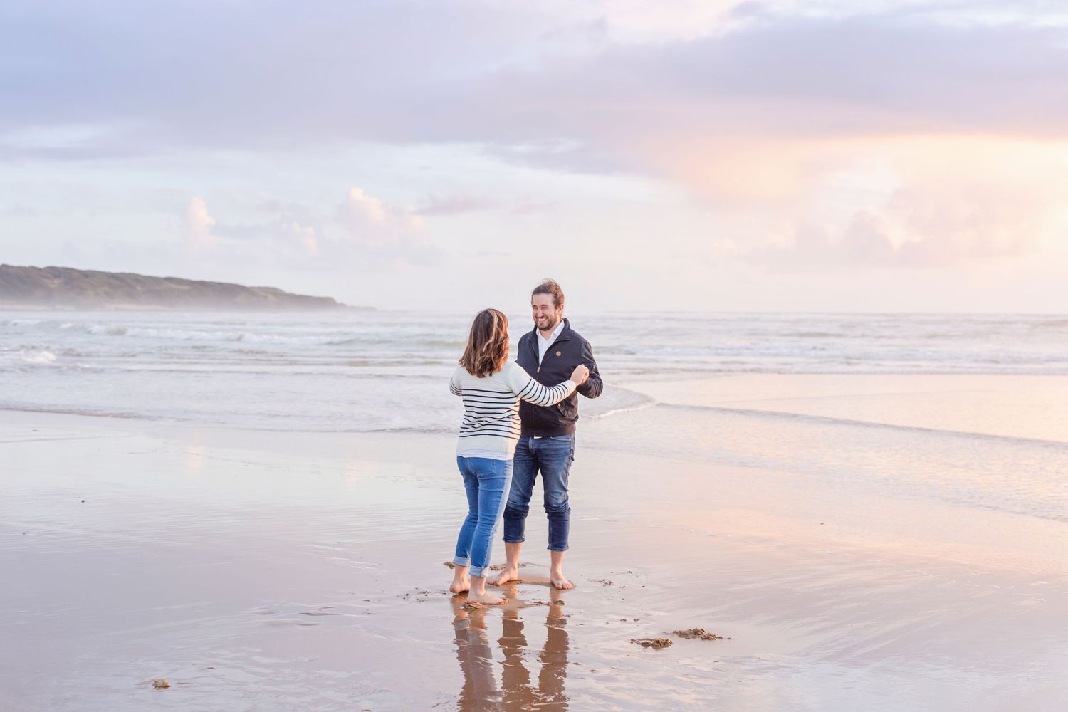 A couple stands together in ankle-deep ocean water during sunset.
