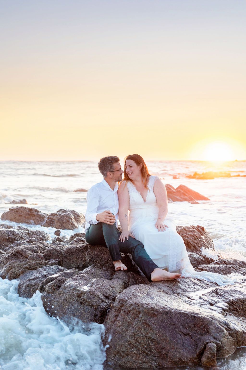 A couple shares a laugh while sitting on ocean rocks at sunset.
