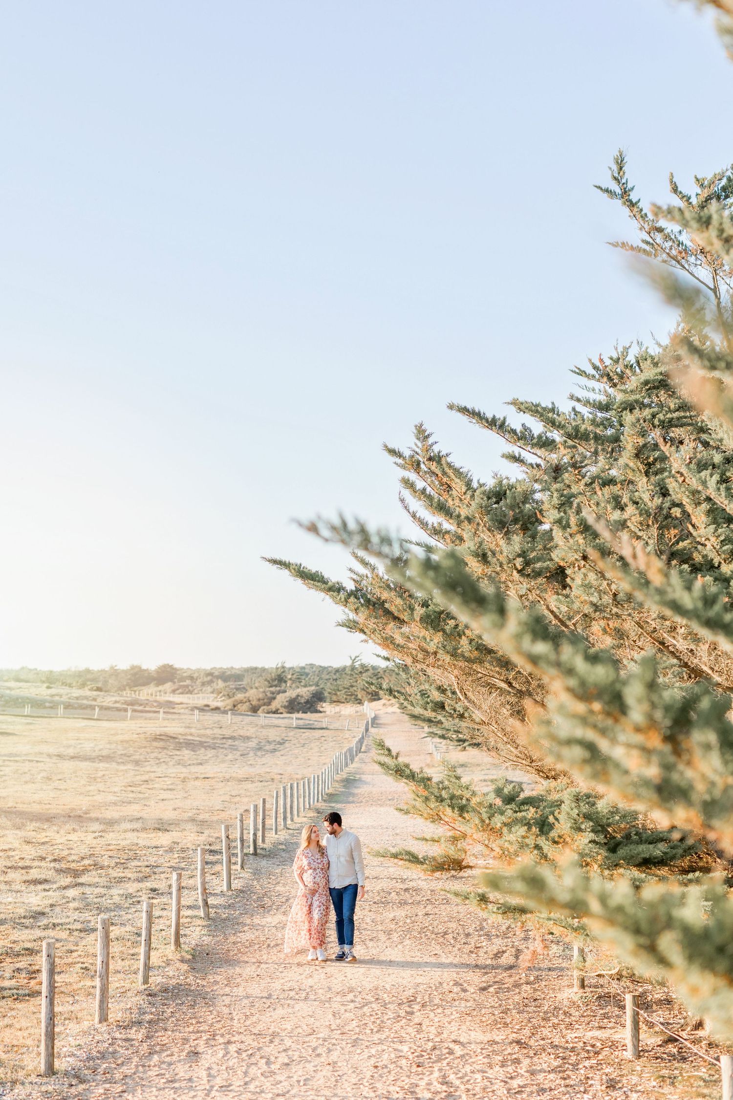 A couple walking down a scenic dirt path lined with trees and fencing at sunset.