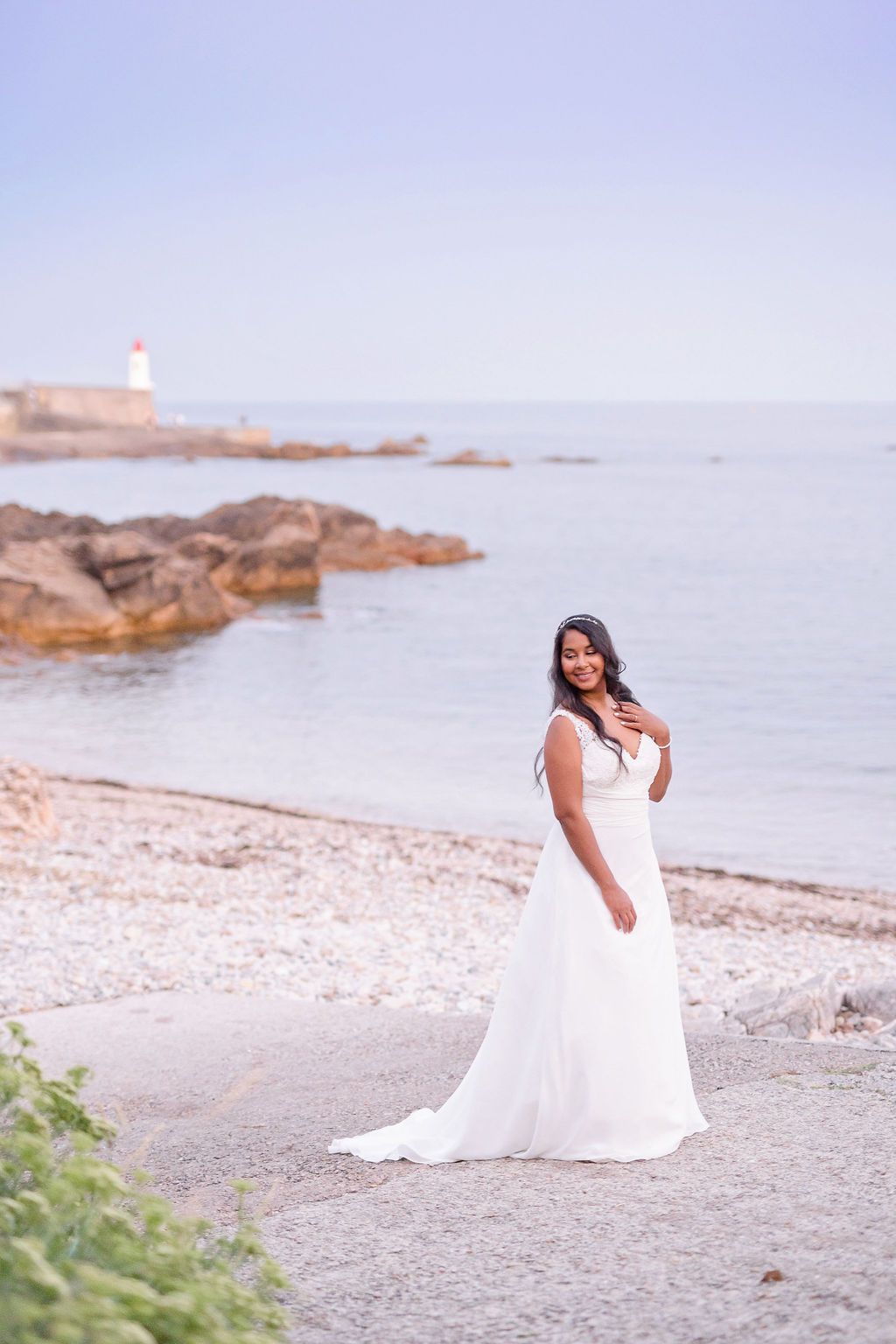 White wedding gown flows across a rocky coastal beach with lighthouse in background.