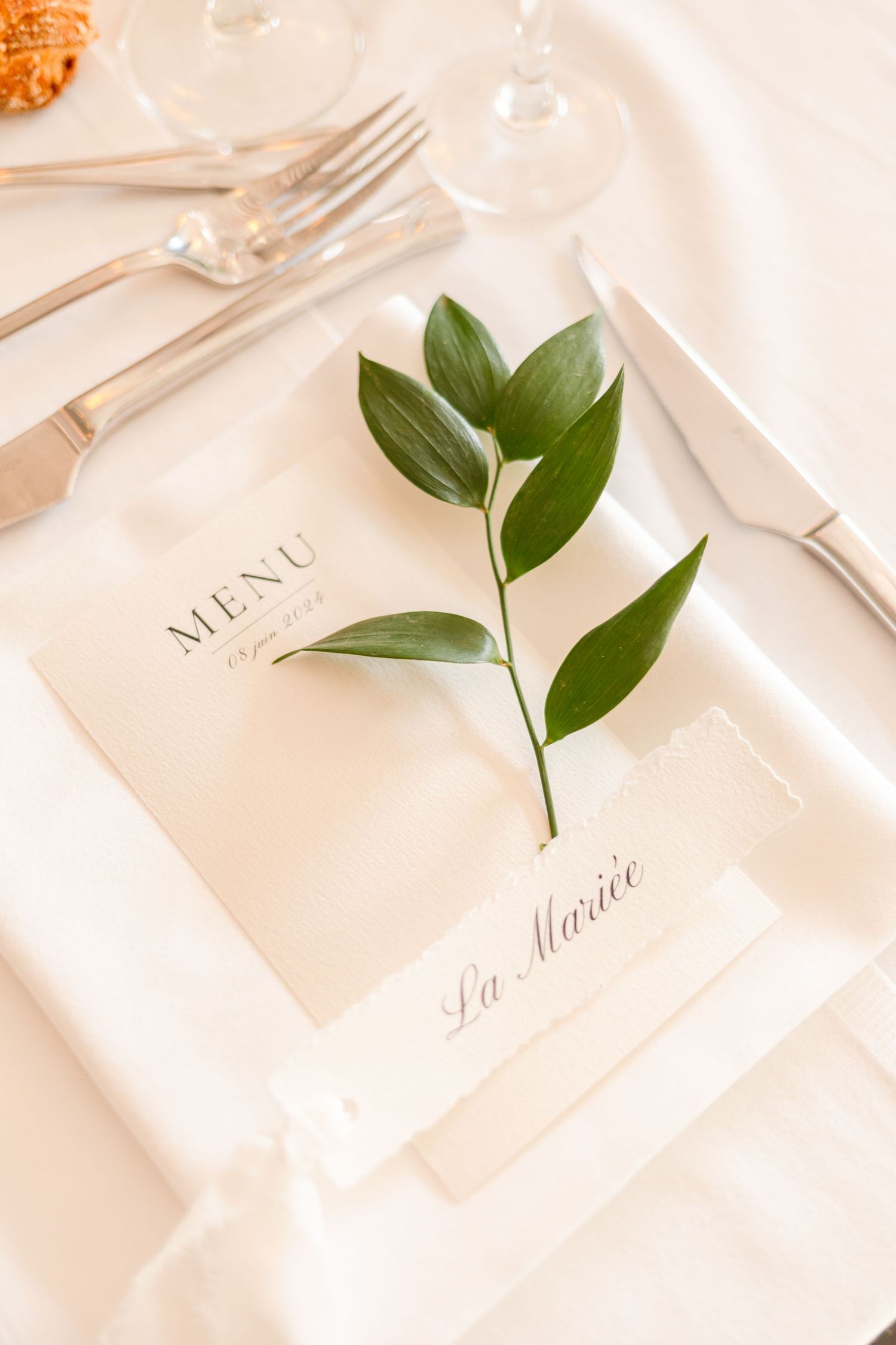 Elegant place setting with menu card and fresh green leaves on white tablecloth.