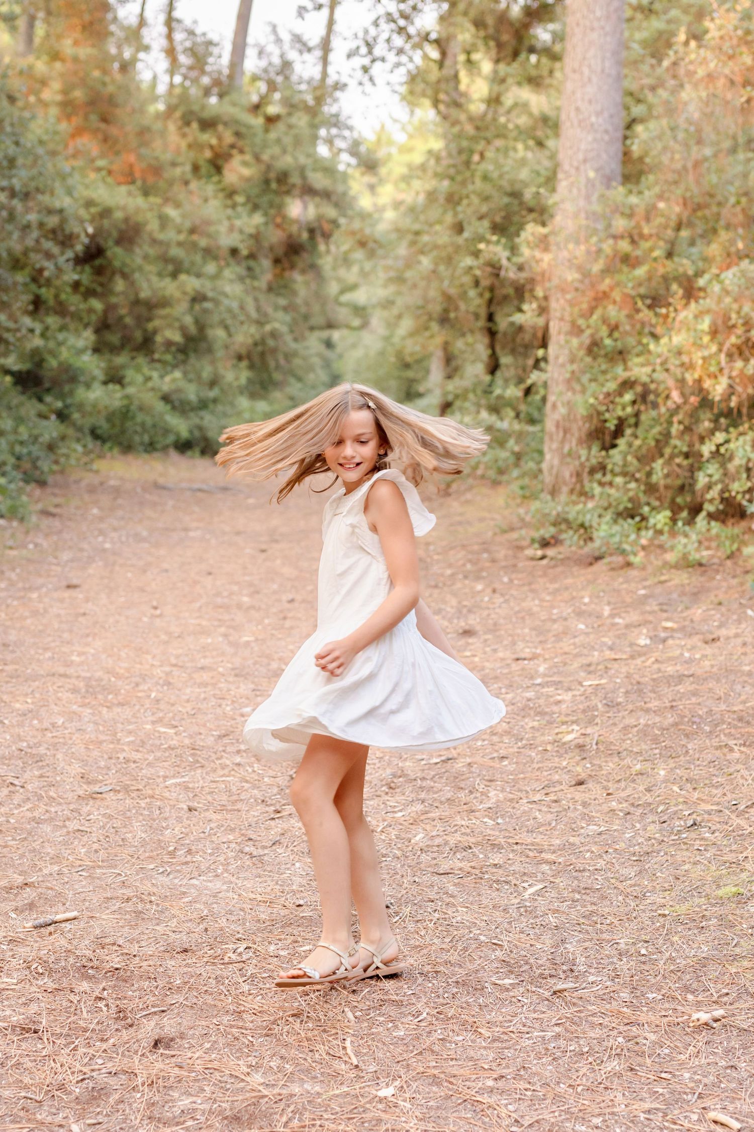 Person in white dress twirling playfully on a forest trail with hair flowing in motion.