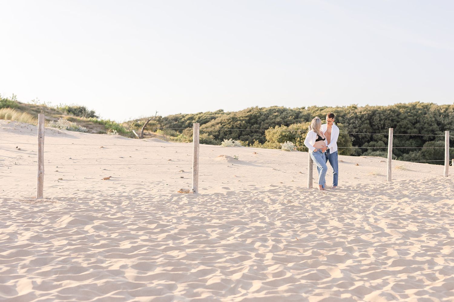 A couple embraces on a serene sandy beach with wooden posts and coastal dunes in the background.