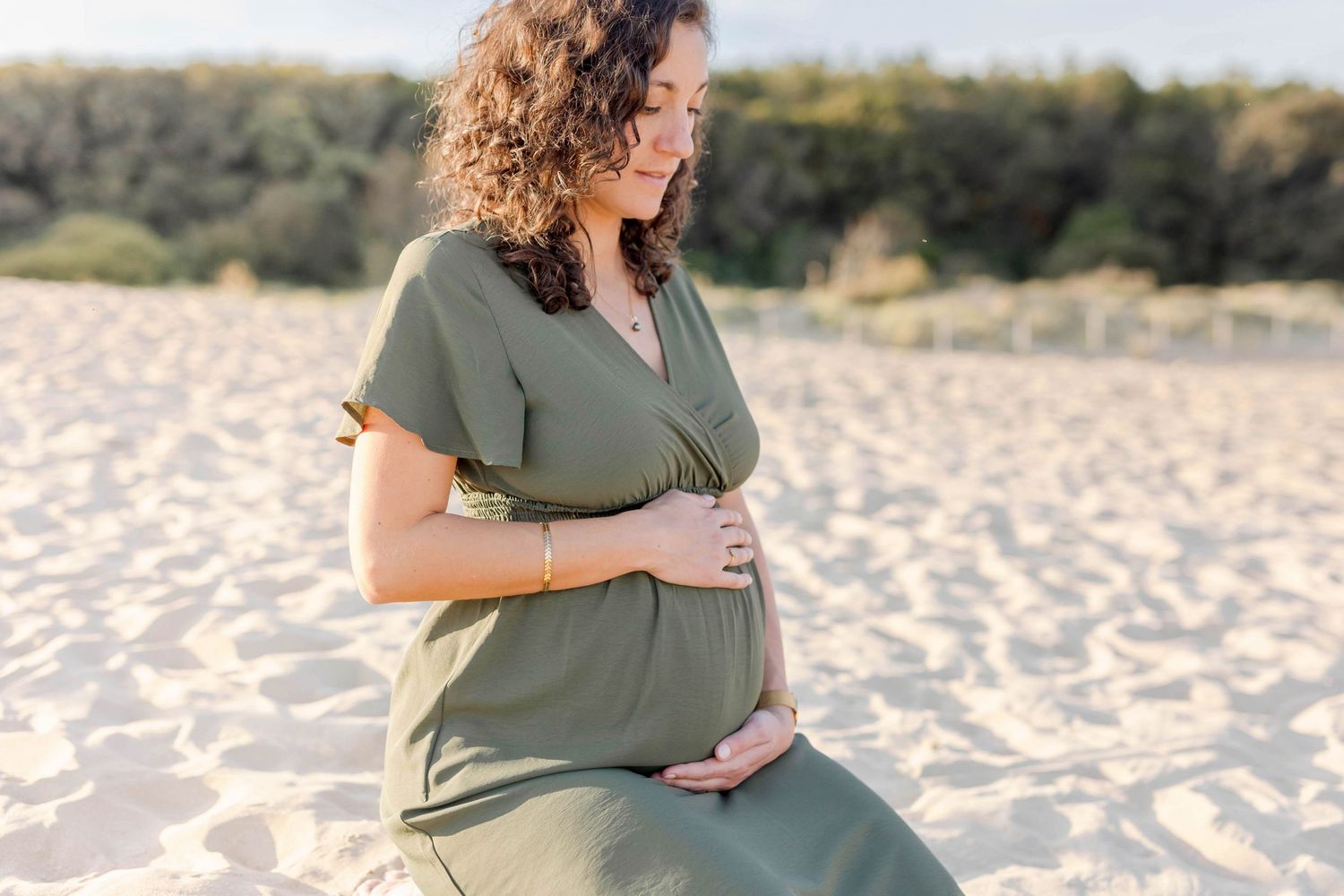 An expectant mother in an olive dress sits peacefully on sandy beach during sunset.