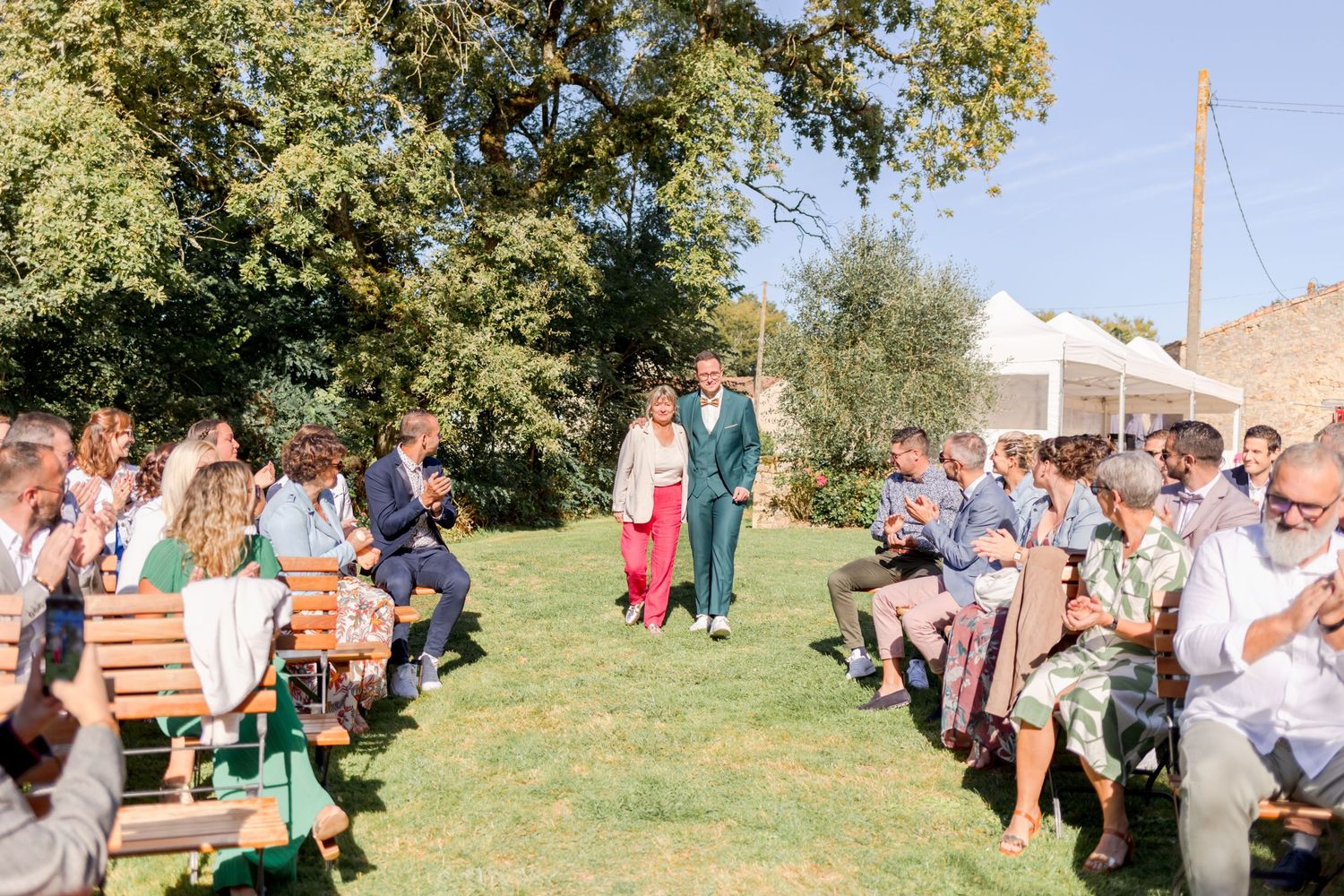 Guests seated on wooden benches at outdoor wedding ceremony under trees.