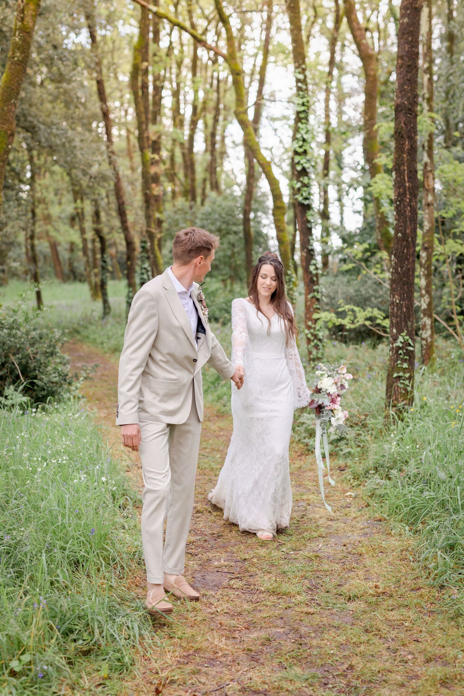 A romantic woodland wedding scene shows a couple in tan suit and white lace dress walking along a forest path.