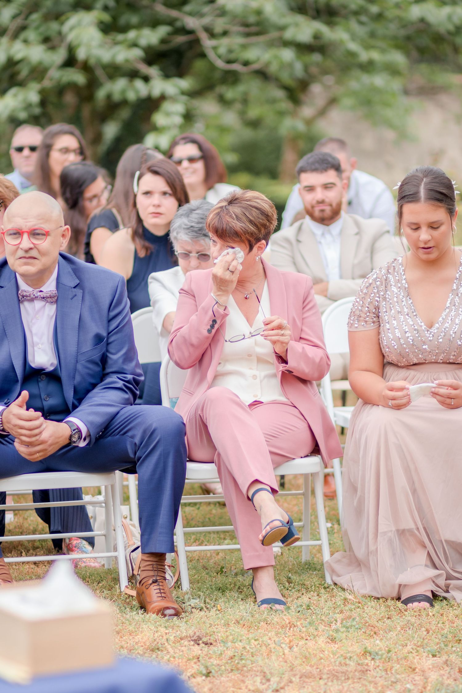 Wedding guests in formal attire seated on white chairs during an outdoor ceremony.