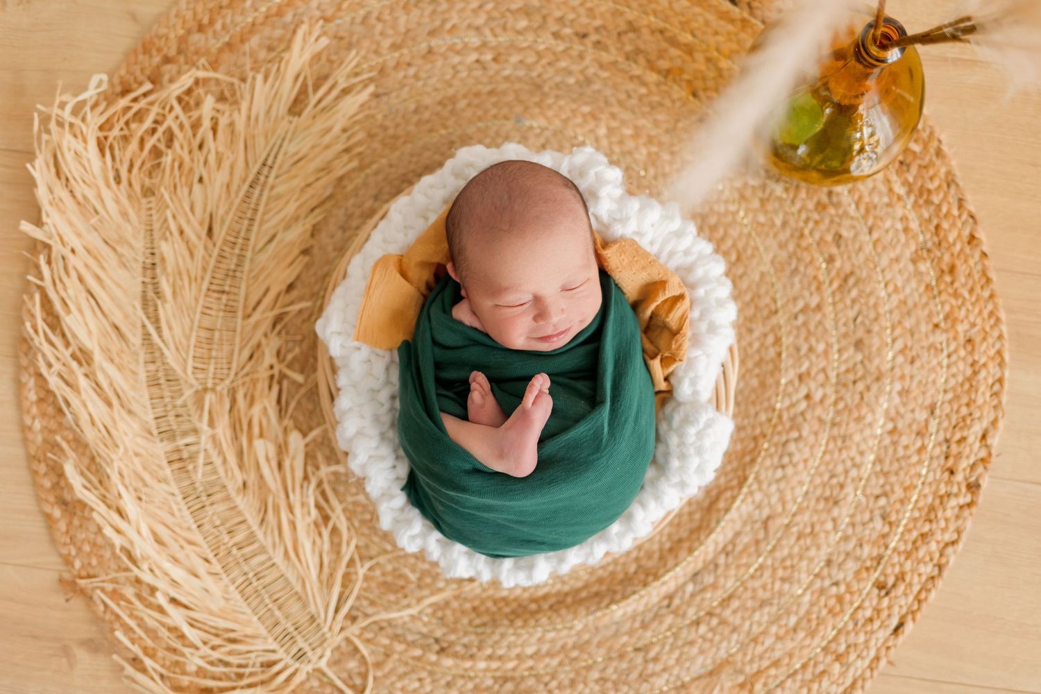 Newborn wrapped in emerald green swaddle sleeping in circular woven basket with white cushioning.