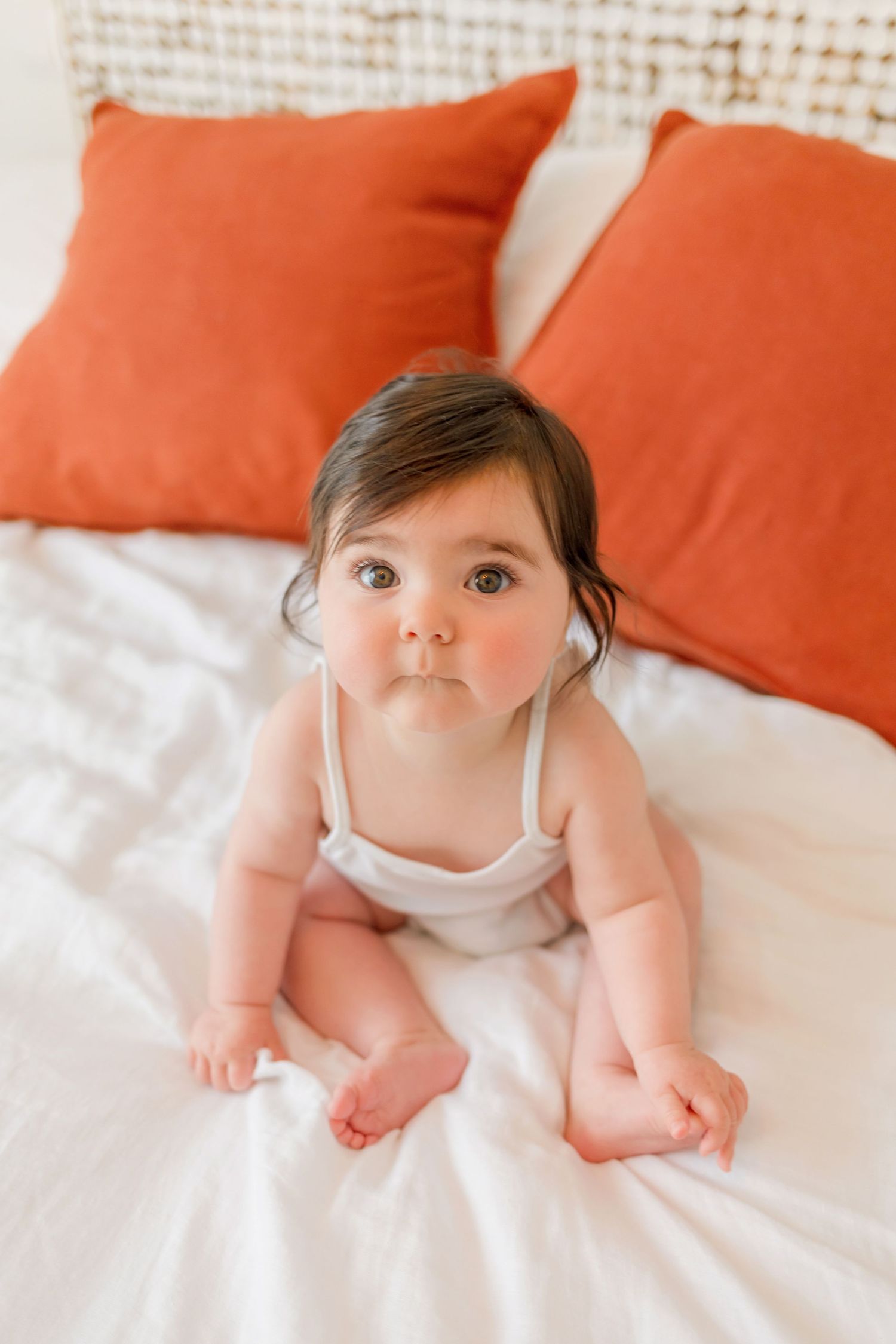 Baby in white outfit lying on bed with orange pillows in bright natural light setting.