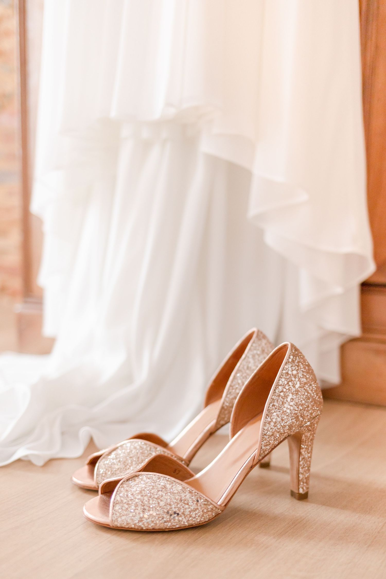 Glittery champagne-colored peep-toe wedding heels against a white dress background.