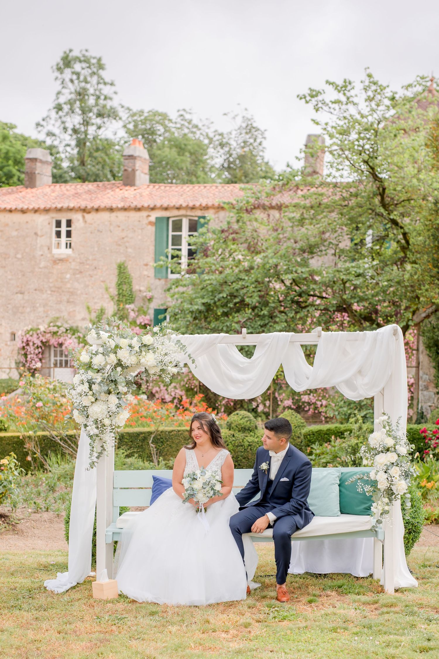A wedding couple sits on a bench under a white draped arch decorated with flowers in a garden setting.