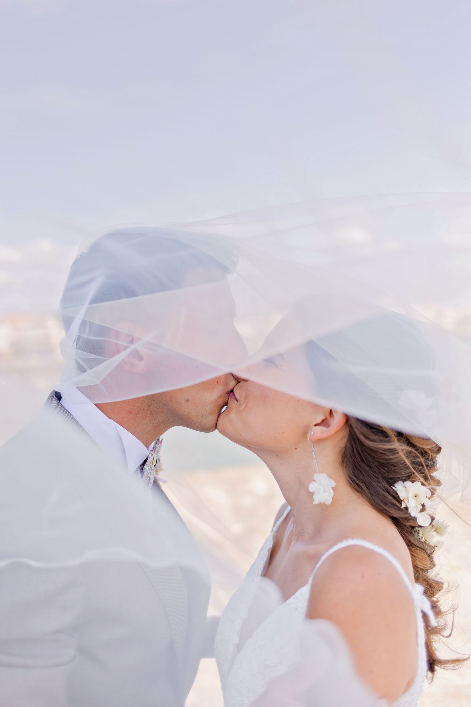 Newlyweds share a tender kiss under flowing veil against bright sky background.
