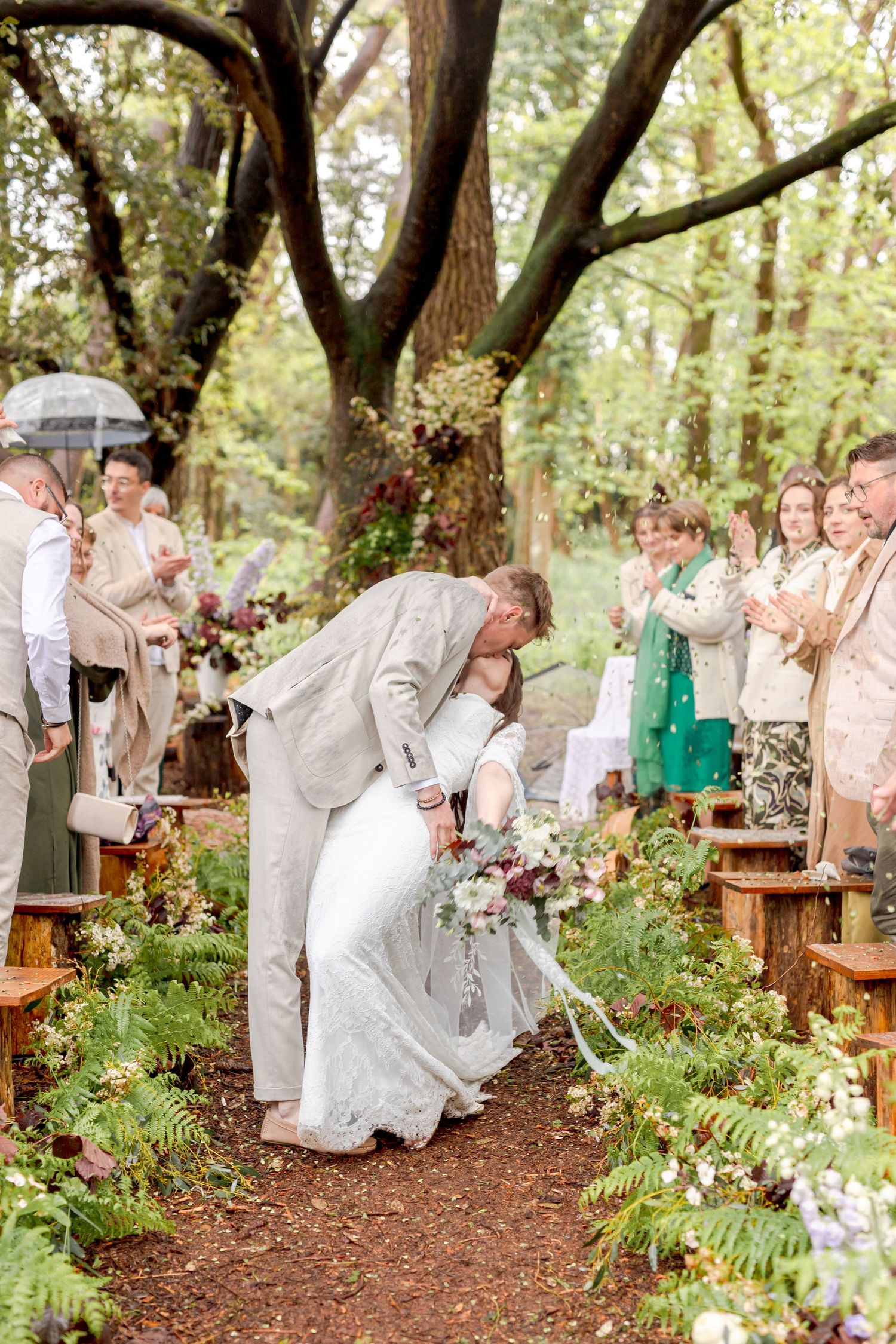 Newlyweds share a dip kiss while guests cheer at a forest wedding ceremony.