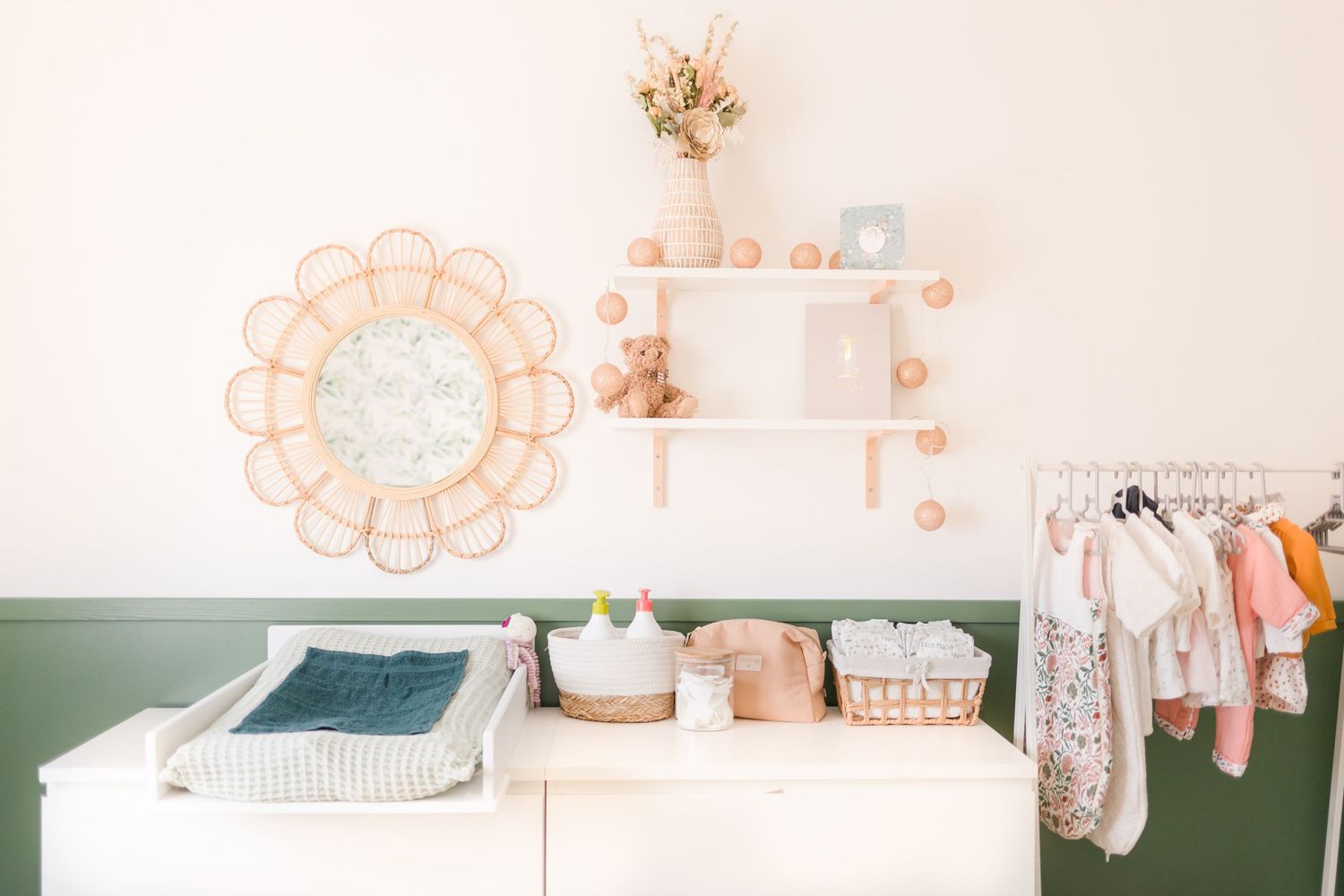 Nursery changing station with rattan mirror, floating shelves, and clothes rack in soft pink and green tones.