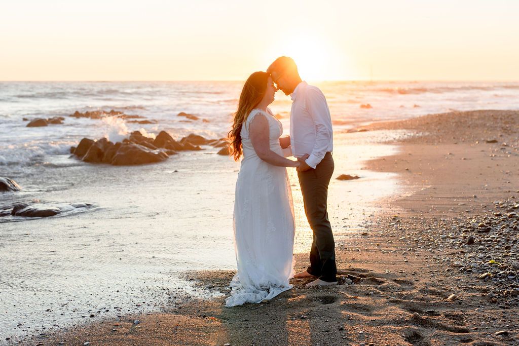 A romantic couple embraces on a beach at sunset with rocky coastline in the background.