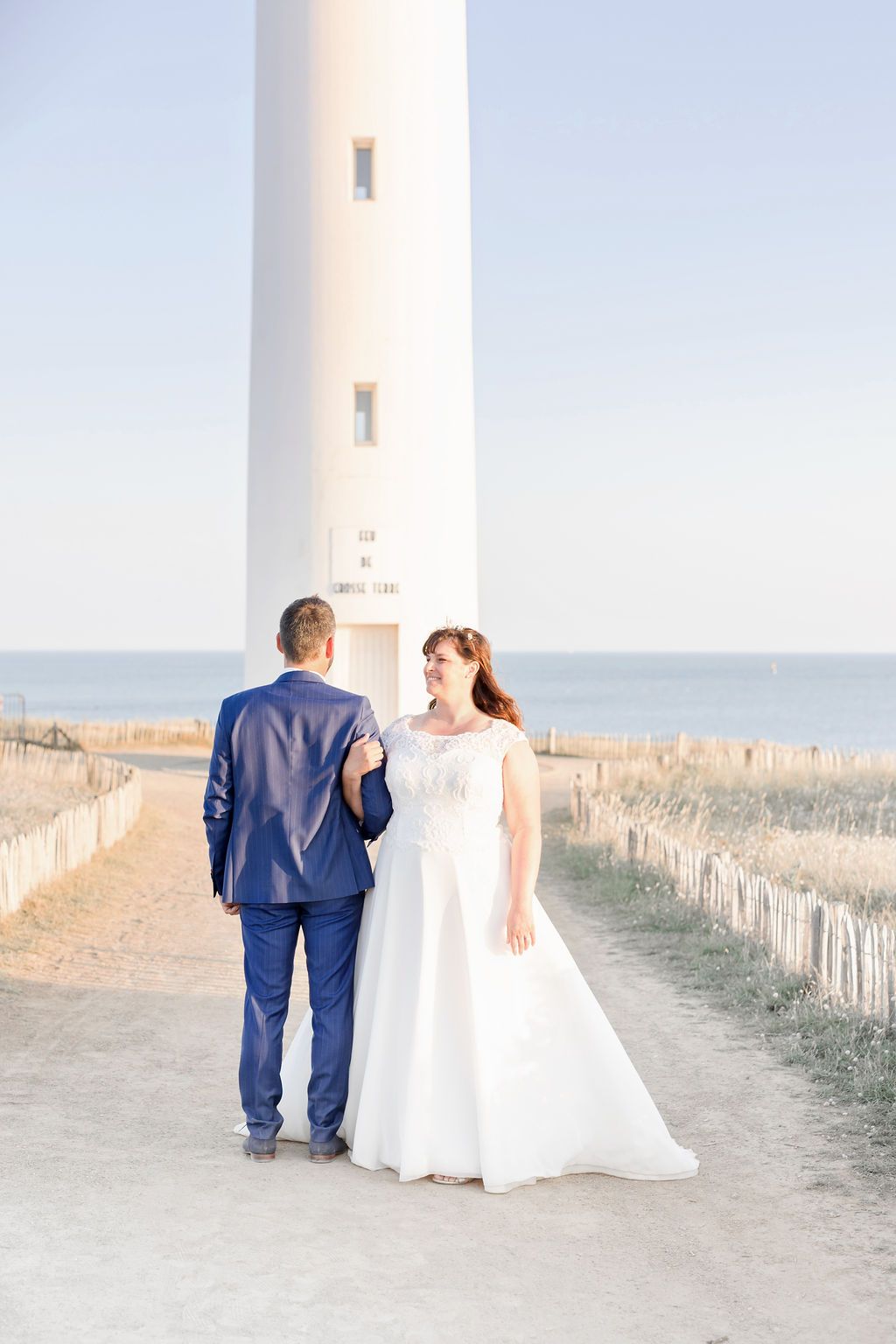 Wedding couple walking along a coastal path beside a tall white lighthouse.