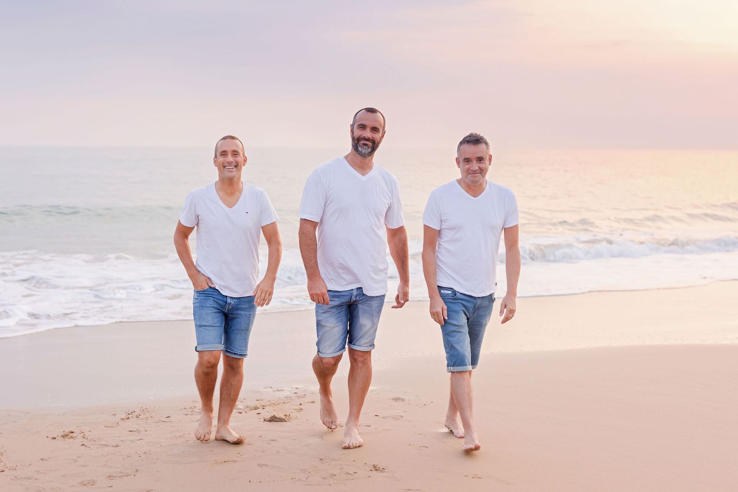 Three people in white t-shirts and denim shorts walking barefoot on beach at sunset.