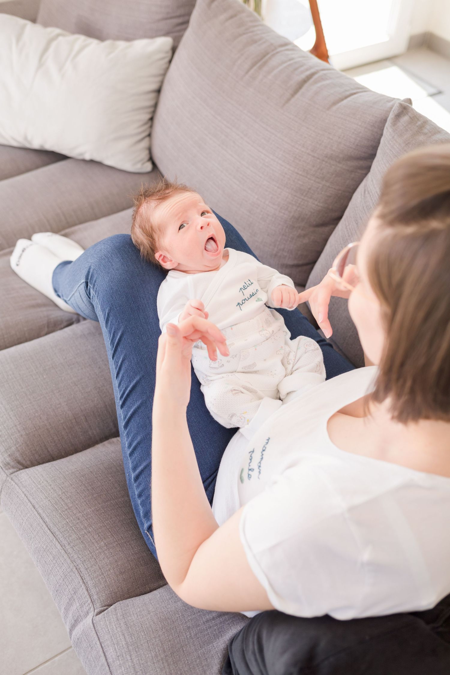 Baby in white onesie smiling and playing while laying on grey couch.