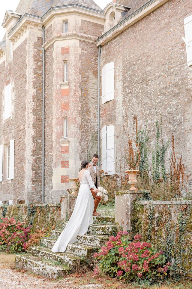 White wedding dress flowing elegantly on stone steps with blooming red flowers against rustic chateau walls.