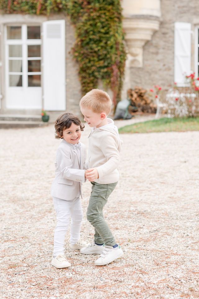Two small children in white outfits playfully interacting on gravel with an ivy-covered building in the background.