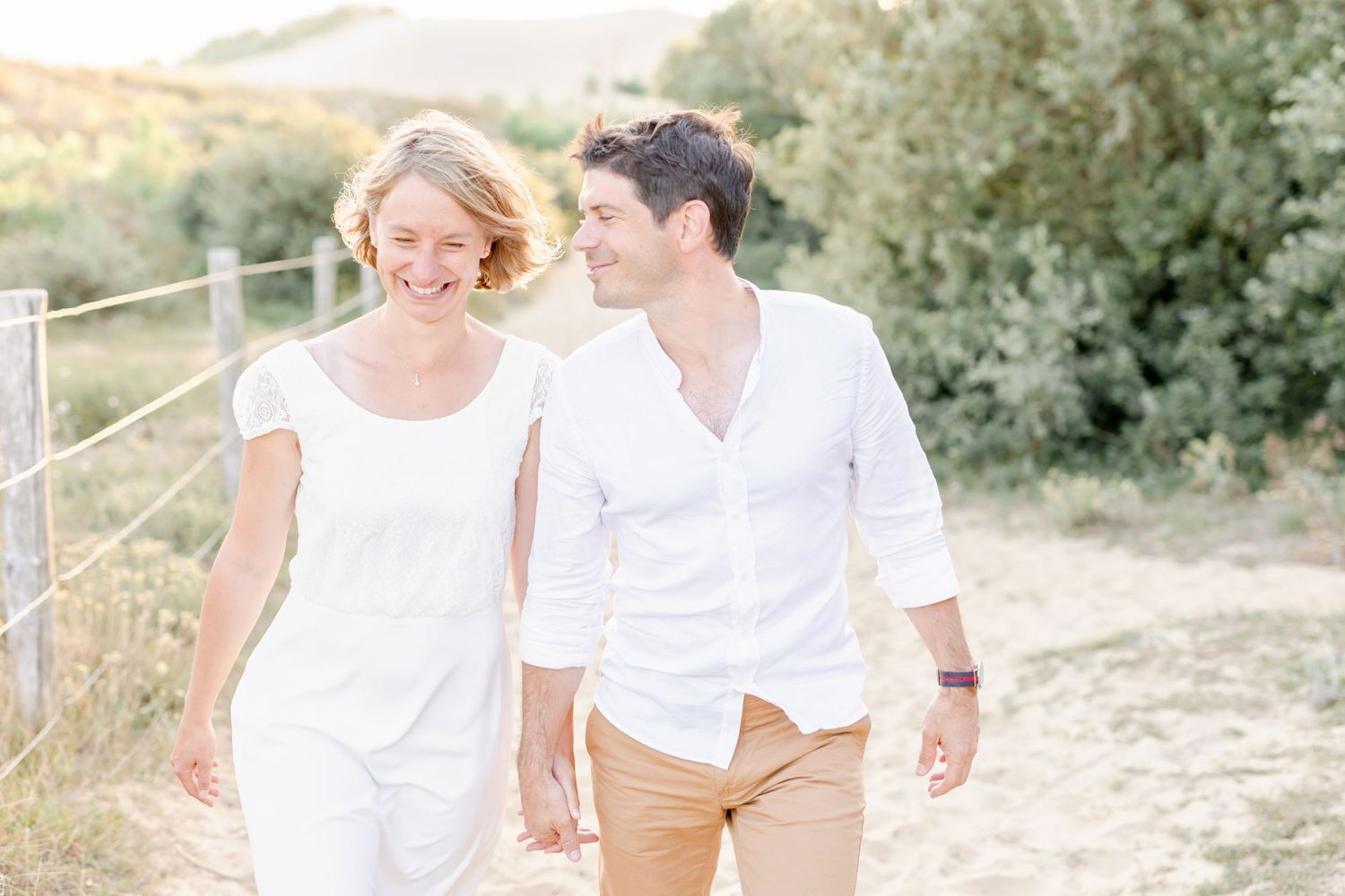 Romantic couple in coordinated white outfits walk hand in hand along a sunlit beach path.
