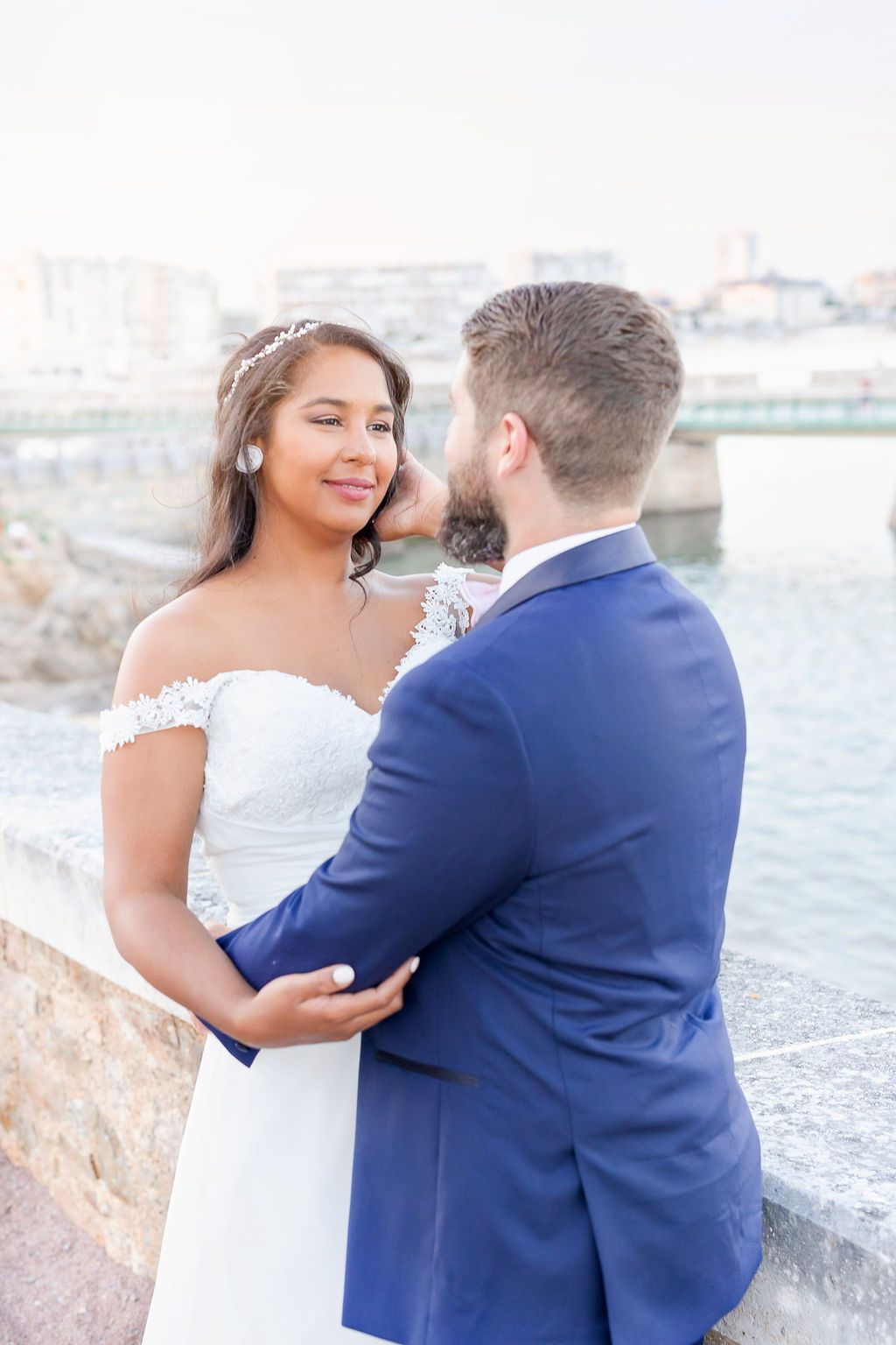 Elegant bride in off-shoulder dress and groom in navy suit share an intimate moment by the water.