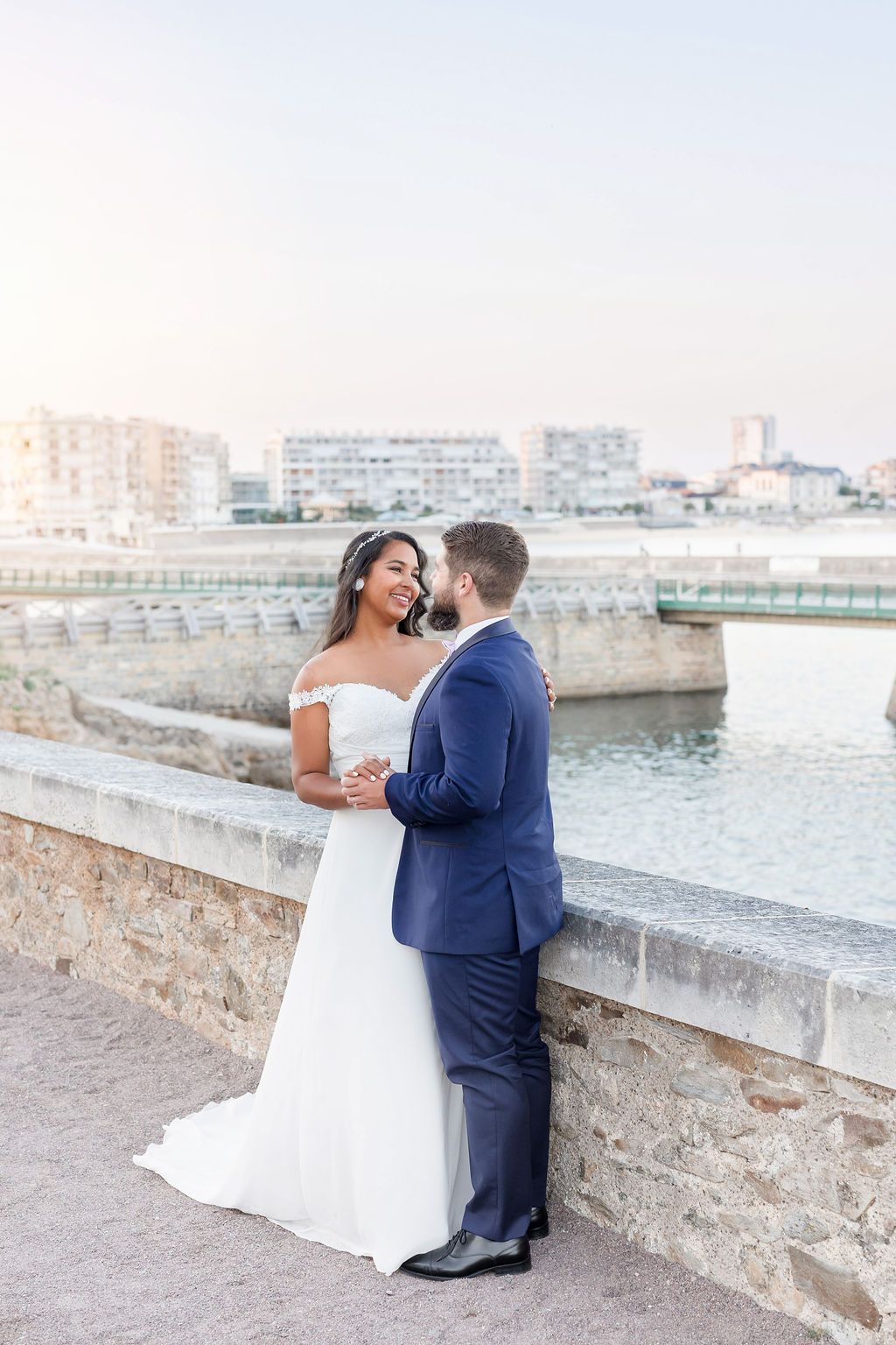 A romantic wedding couple embraces on a stone bridge overlooking the water during sunset.