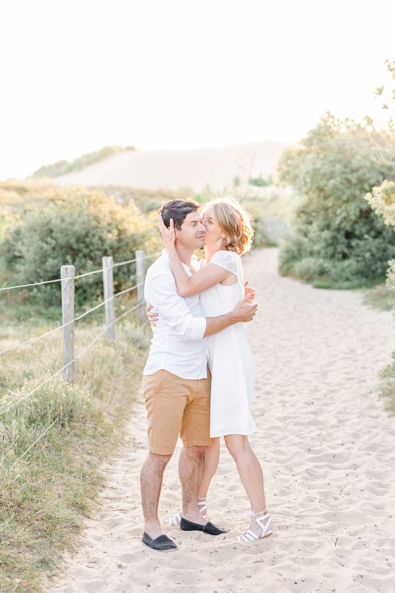A couple in white and khaki outfits shares a tender moment on sandy beach path during golden hour.