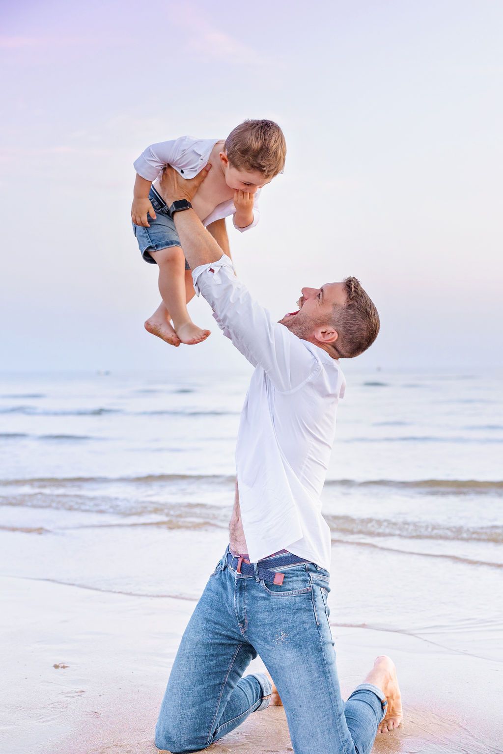 A person in a white shirt and jeans lifts a small child playfully on the beach at sunset.