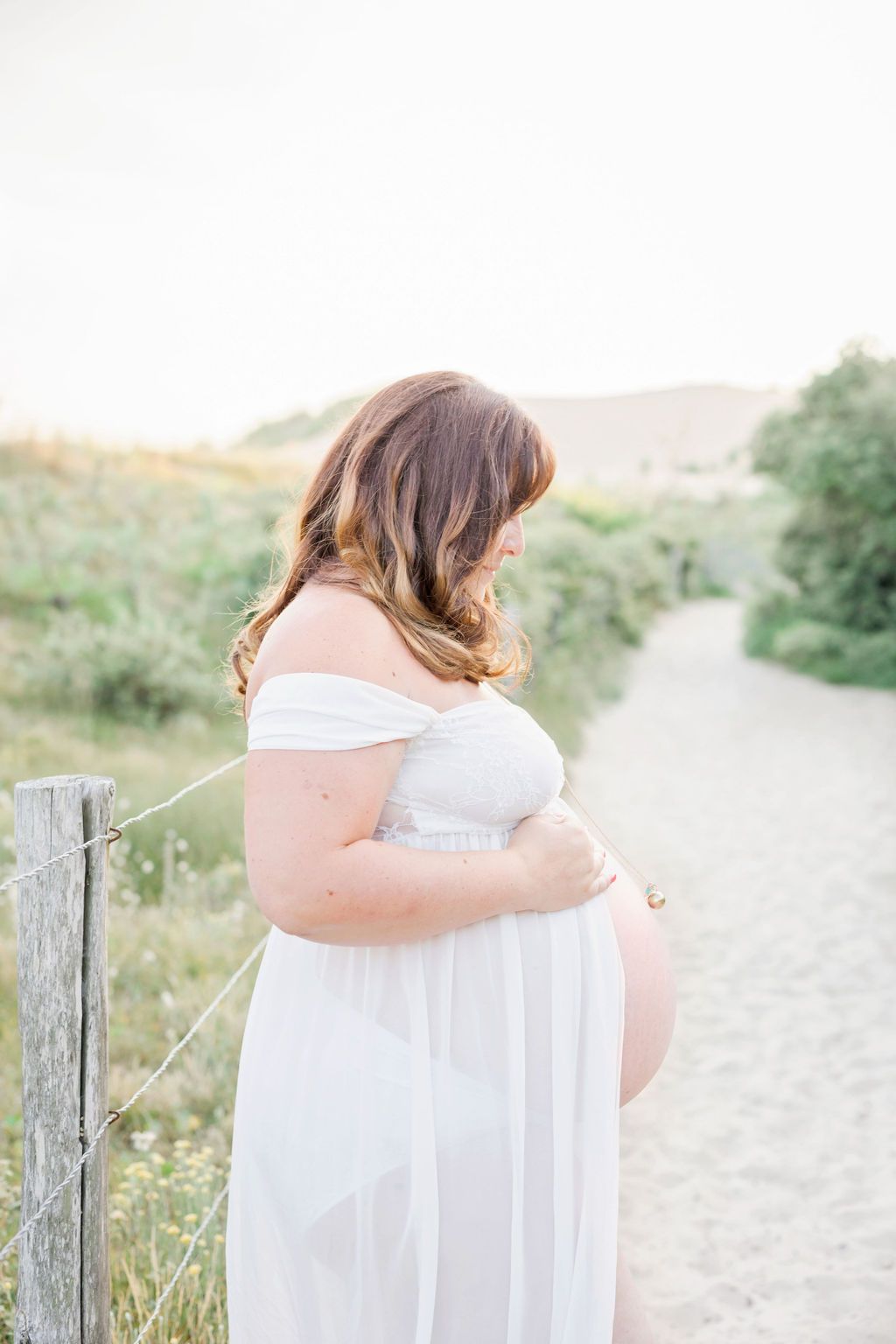 Woman in ethereal white dress standing on sandy path with coastal greenery.