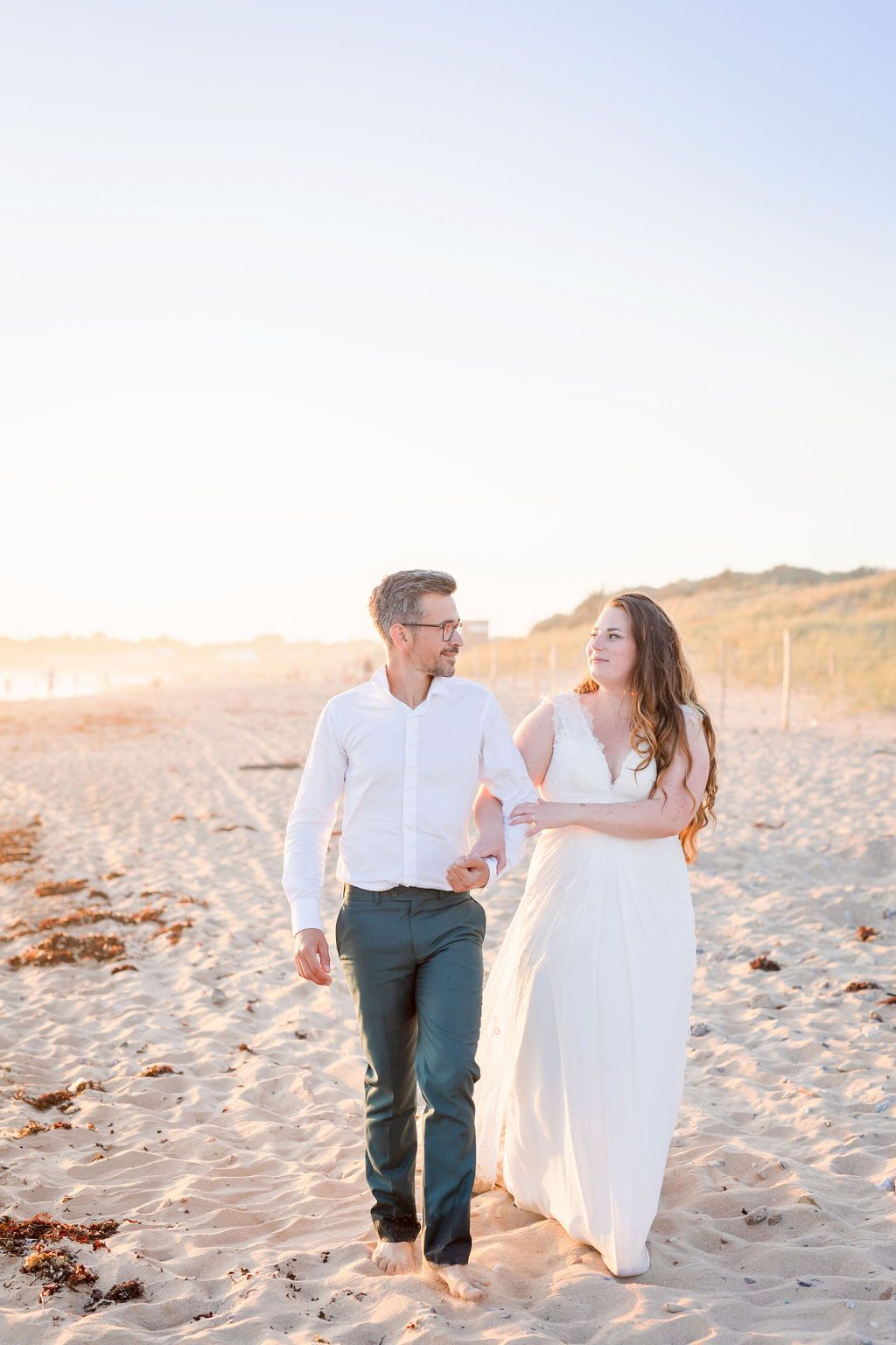 A couple walks hand in hand on a sandy beach at sunset wearing white and earth-toned clothing.