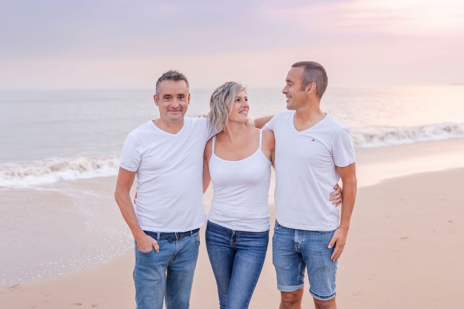 Three people in coordinated white and denim outfits pose on beach at sunset.